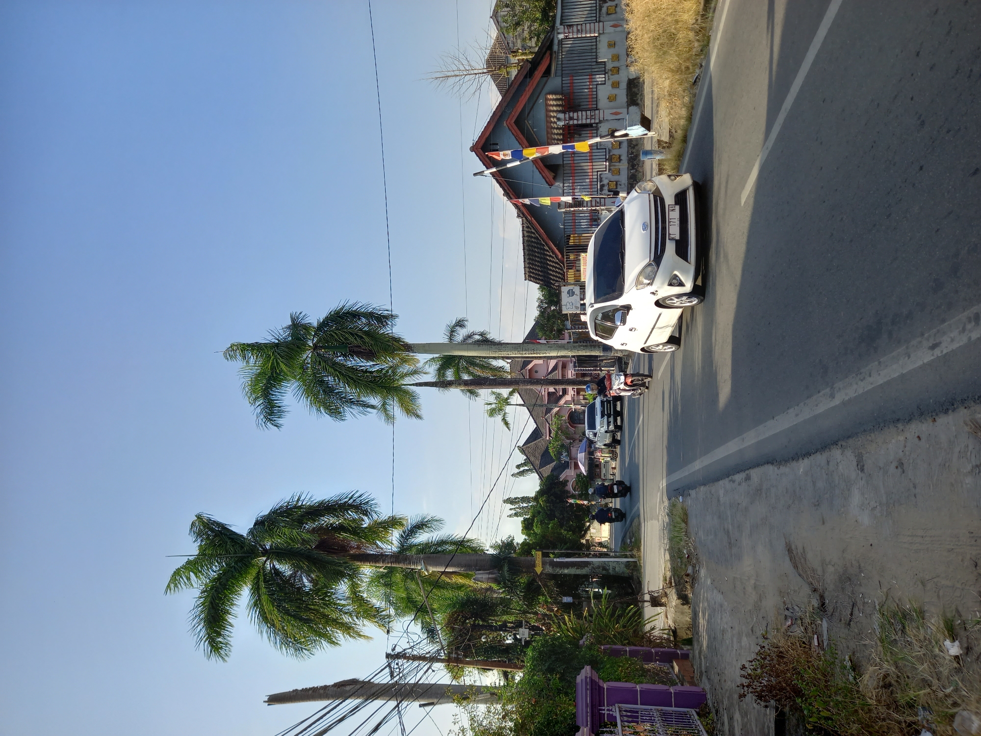 The image captures a street scene in Samarinda, Indonesia, under a clear blue sky. In the foreground, a white car with license plate KT 171 NA is driving on an asphalt road with a white dividing line. To the left of the road, there is a sidewalk with a purple fence and some vegetation. Tall palm trees line the street, casting shadows on the road. Several motorcycles are also visible on the road, with riders wearing helmets. In the background, houses with red roofs and gray walls are visible, some decorated with colorful flags. The environment appears to be a residential area with some commercial activity, as suggested by a sign on a pole indicating "DIJUAL" (For Sale). The overall atmosphere is that of a sunny day in a tropical city.