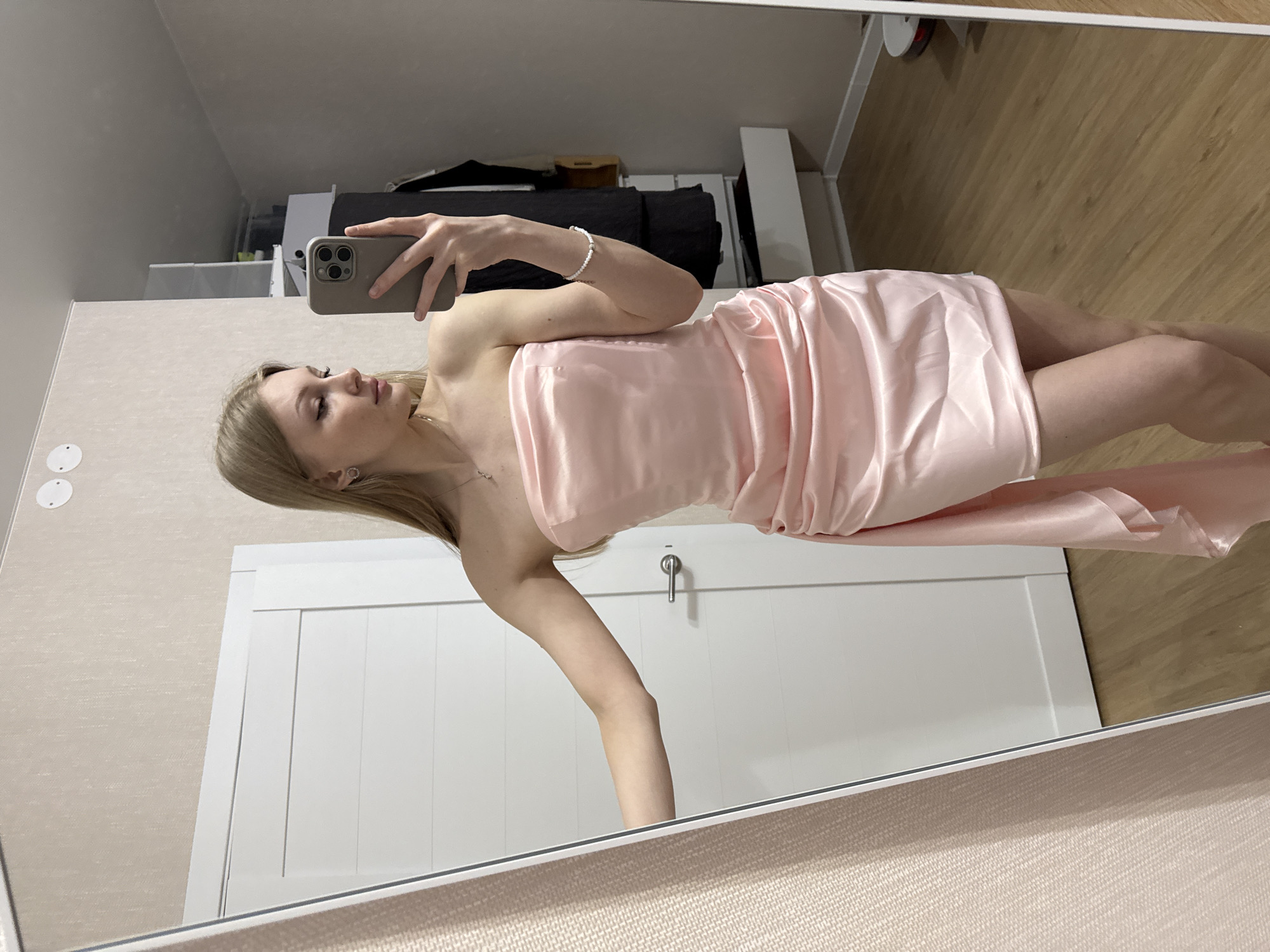 A young woman is reflected in a mirror, taking a selfie. She has blonde hair and is wearing a light pink dress that appears to be satin. She is holding a smartphone in her left hand. The background includes a white door with a silver handle, a light-colored wall, a wooden floor, and a black cushioned object on top of a white piece of furniture. The location appears to be inside a room. The location, Perm, Russia, is not verifiable in the image.