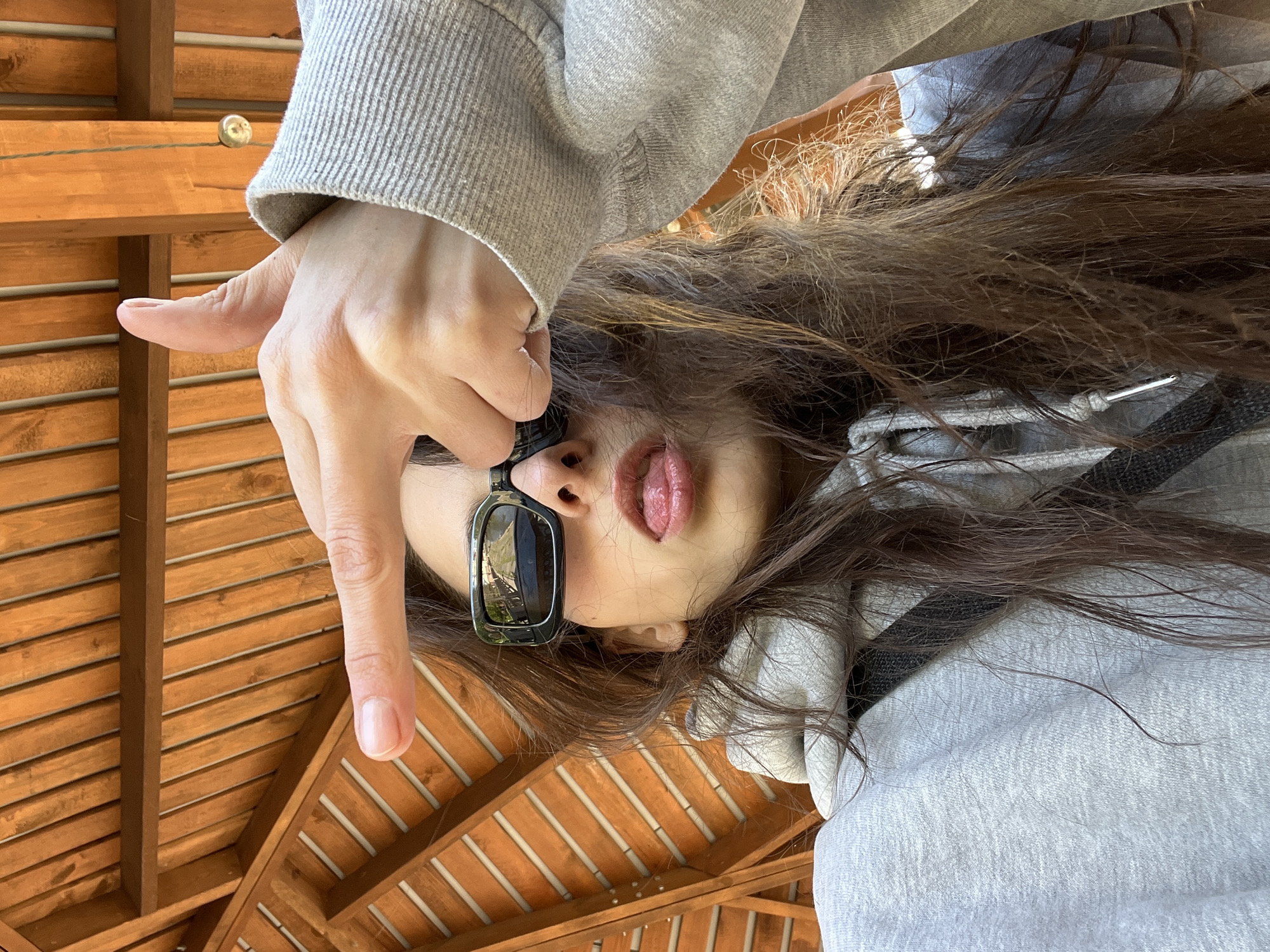 The image shows a woman with long brown hair and wearing sunglasses, viewed from below. Her tongue is sticking out. She is wearing a gray hooded sweatshirt. Her right hand, with the index finger extended, is visible near the top left of the frame. The background reveals a wooden structure with parallel slats, likely a pergola or awning. Based on available information, this photo might be taken from Krasnoyarsk, Russia as an assumption.