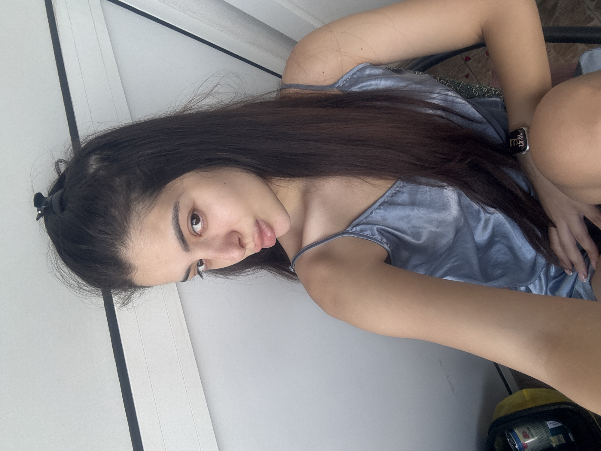 The image shows a young woman with long, dark brown hair looking directly at the camera with a neutral expression. She is angled diagonally, wearing a light blue or silver satin-like top with thin straps. Her hair is partially pulled back with a black clip. On her left wrist, she wears a smartwatch that displays the time "19:42".
The scene is set indoors against a light-colored wall, which features a prominent dark vertical line, possibly indicating a doorframe or paneling. The background also includes a glimpse of a tiled floor and, in the lower right corner, parts of a yellow bag and a silver can are faintly visible. The overall impression is a casual, indoor environment. The location context provided for this image is Plovdiv, Bulgaria.
