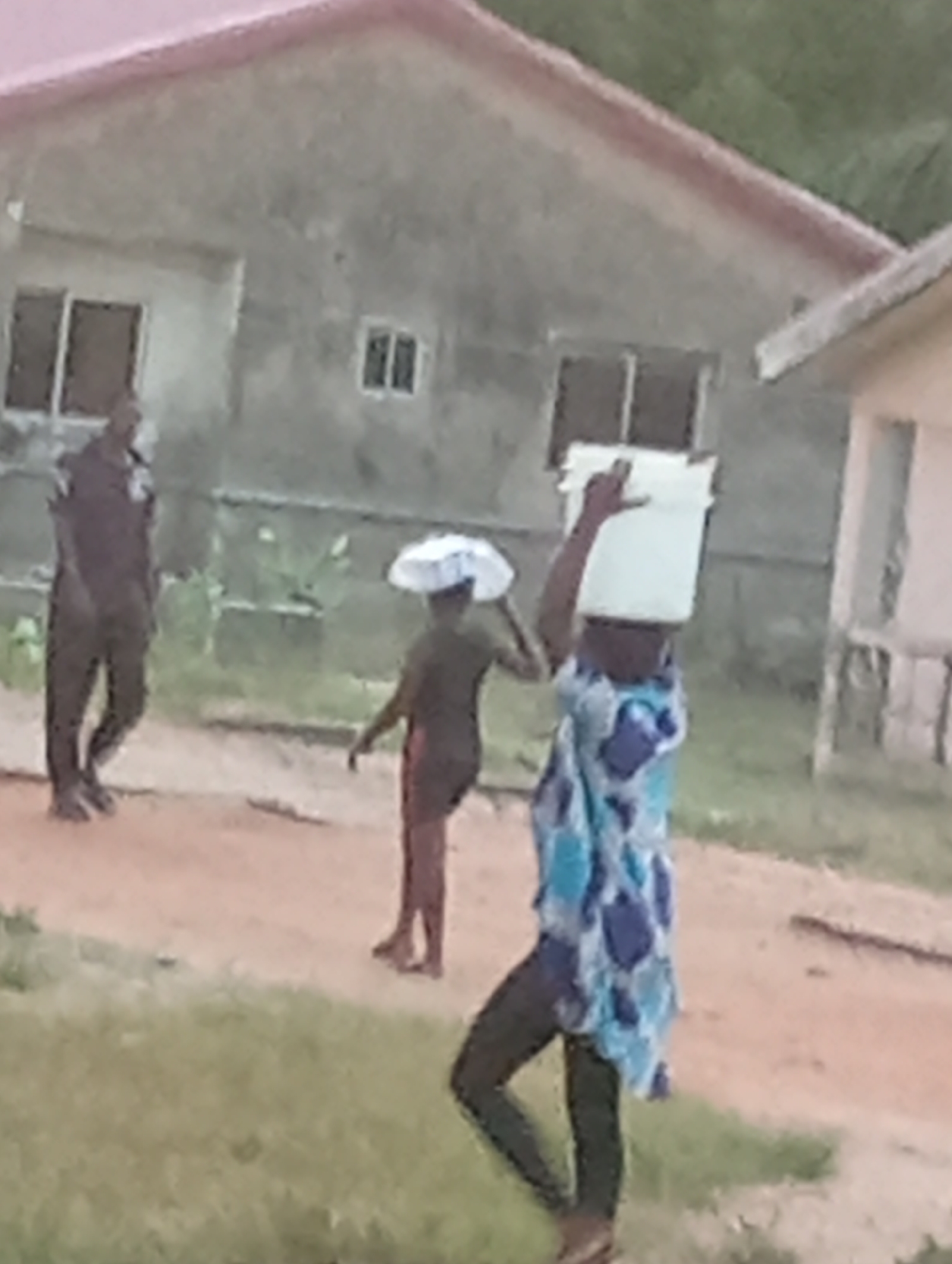 The image depicts a scene in an undetermined city or region. In the foreground, a person with a bucket balanced on their head is partially visible, wearing dark pants and a blue patterned shirt. To the left of them, another person is walking with what appears to be a white object or cloth on their head. Farther to the left stands an individual in what appears to be a dark uniform. Behind them is a building, and a second, smaller building is to the right of the frame. The ground consists of a mix of grassy areas and what appears to be bare earth.
