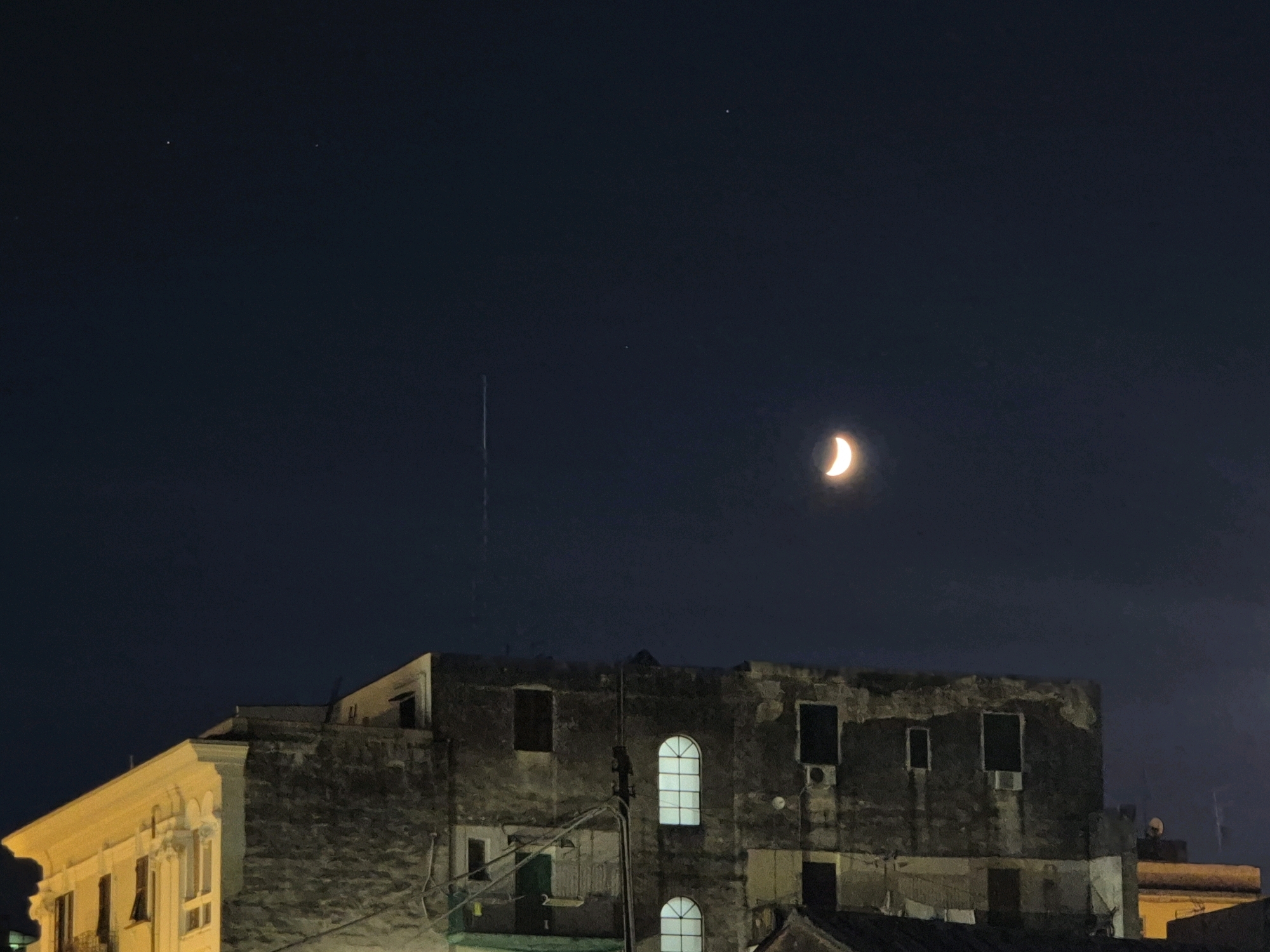 The image captures a crescent moon in a dark, starry sky above a city. Below the sky are two buildings, one a lighter colored, ornately decorated structure, and the other a darker, more weathered building with visible signs of disrepair, such as peeling paint and visible air conditioning units. Several windows are visible on both buildings, some illuminated from within. The overall scene suggests an urban environment at night. The location is identified as Taranto, Italy.