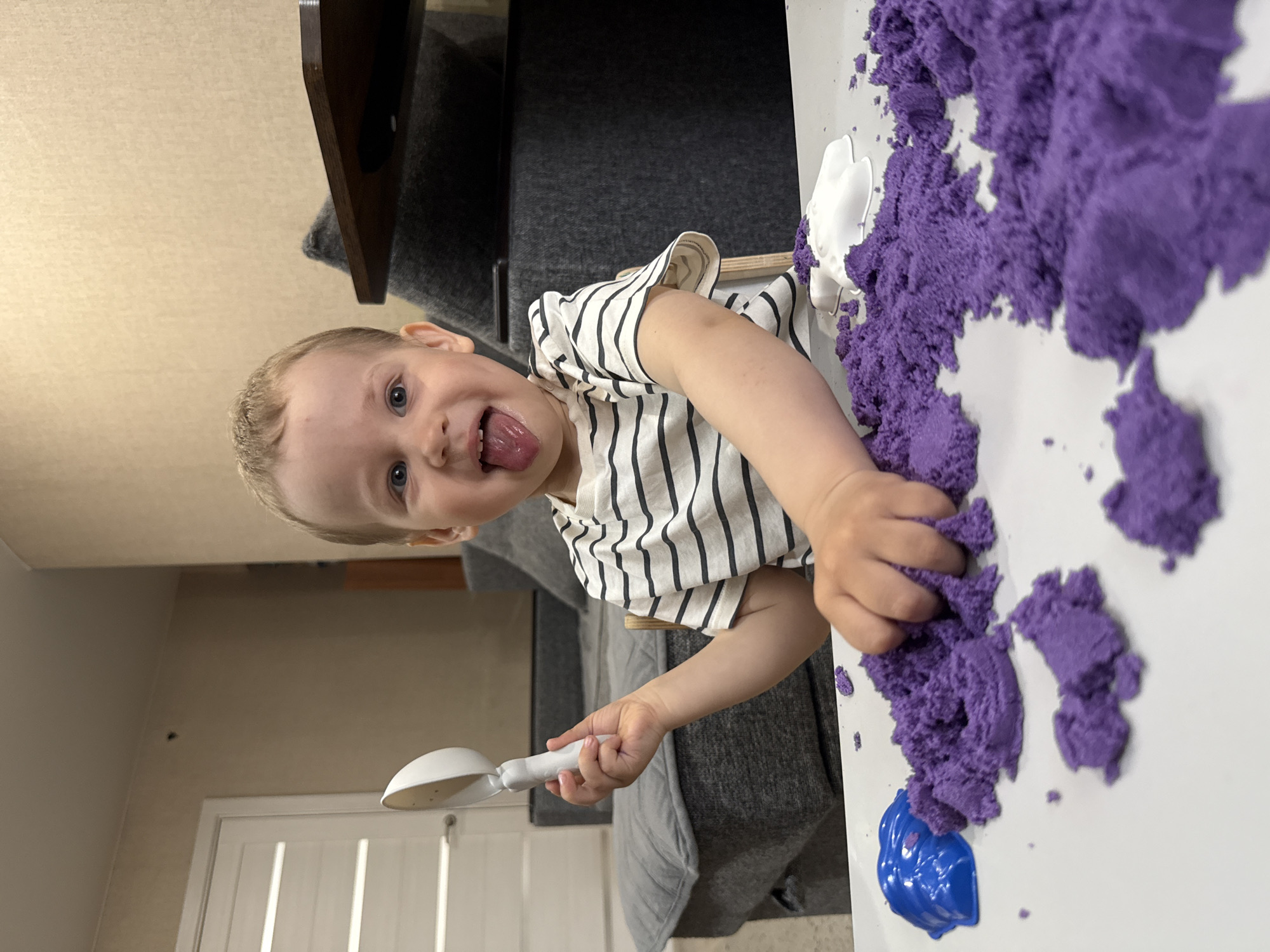 A child with blond hair, wearing a striped shirt, is playing with purple kinetic sand on a white surface. The child is holding a white plastic scoop and sticking their tongue out. In the background, there is a gray couch, a wooden table leg, a wall with a white door, and what appears to be part of an interior room. The location is potentially Шебунино, Russia. A blue plastic mold is visible near the sand.