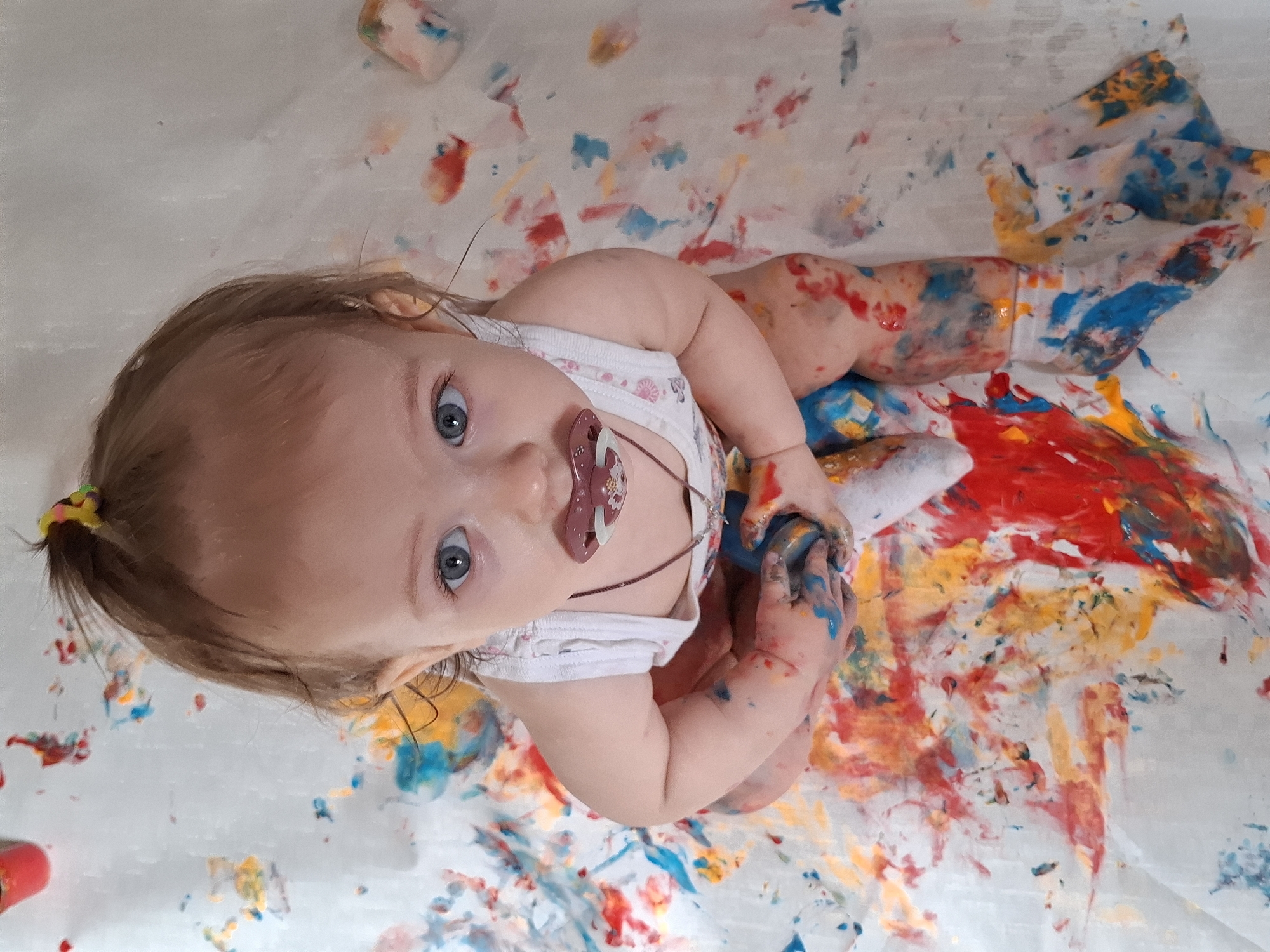 A baby lies on a white surface, covered in multicolored paint. The baby wears a white onesie and has a pacifier in its mouth. The baby's hair is pulled back in a ponytail with a yellow and black hair tie. Paint covers the baby's hands, arms, legs, and the surface around it. The paint is red, blue, yellow, and orange. The location is not specified, so I can't determine if this photo was taken in the city of Chaikovsky, Russia.