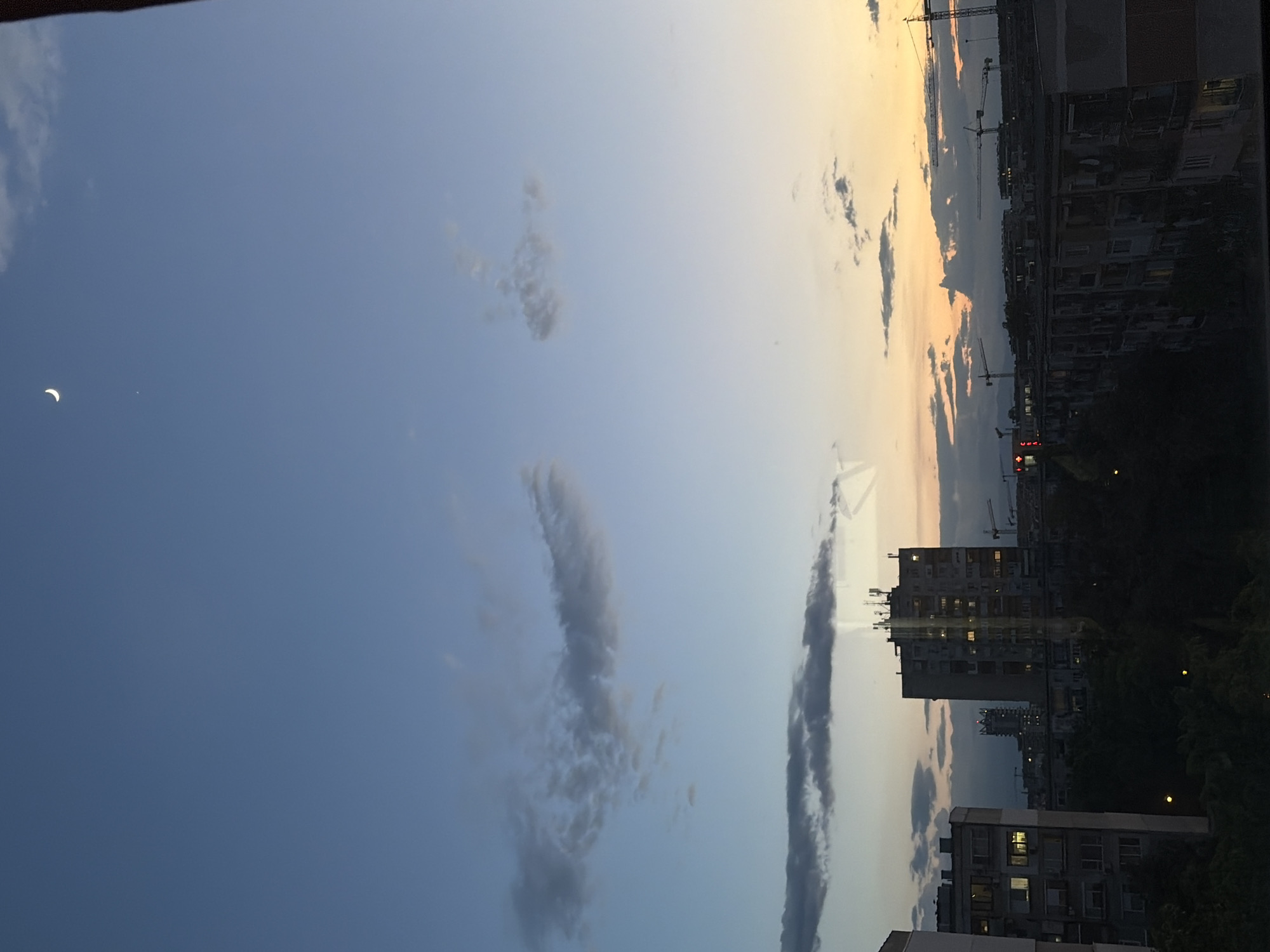 The image captures a cityscape under a twilight sky. The sky transitions from a deep blue on the left to a warm orange and pale yellow on the right horizon, indicating either sunset or sunrise. Clouds of varying density are scattered across the sky. A crescent moon is visible in the upper left corner.
In the foreground, a row of tall residential buildings is visible, some of which are illuminated with interior lights, suggesting it's evening or early morning. Construction cranes punctuate the skyline. Dense vegetation is present at the base of the buildings. The location is most likely Plovdiv, Bulgaria, based on the style of the buildings. No people are visible in the image.