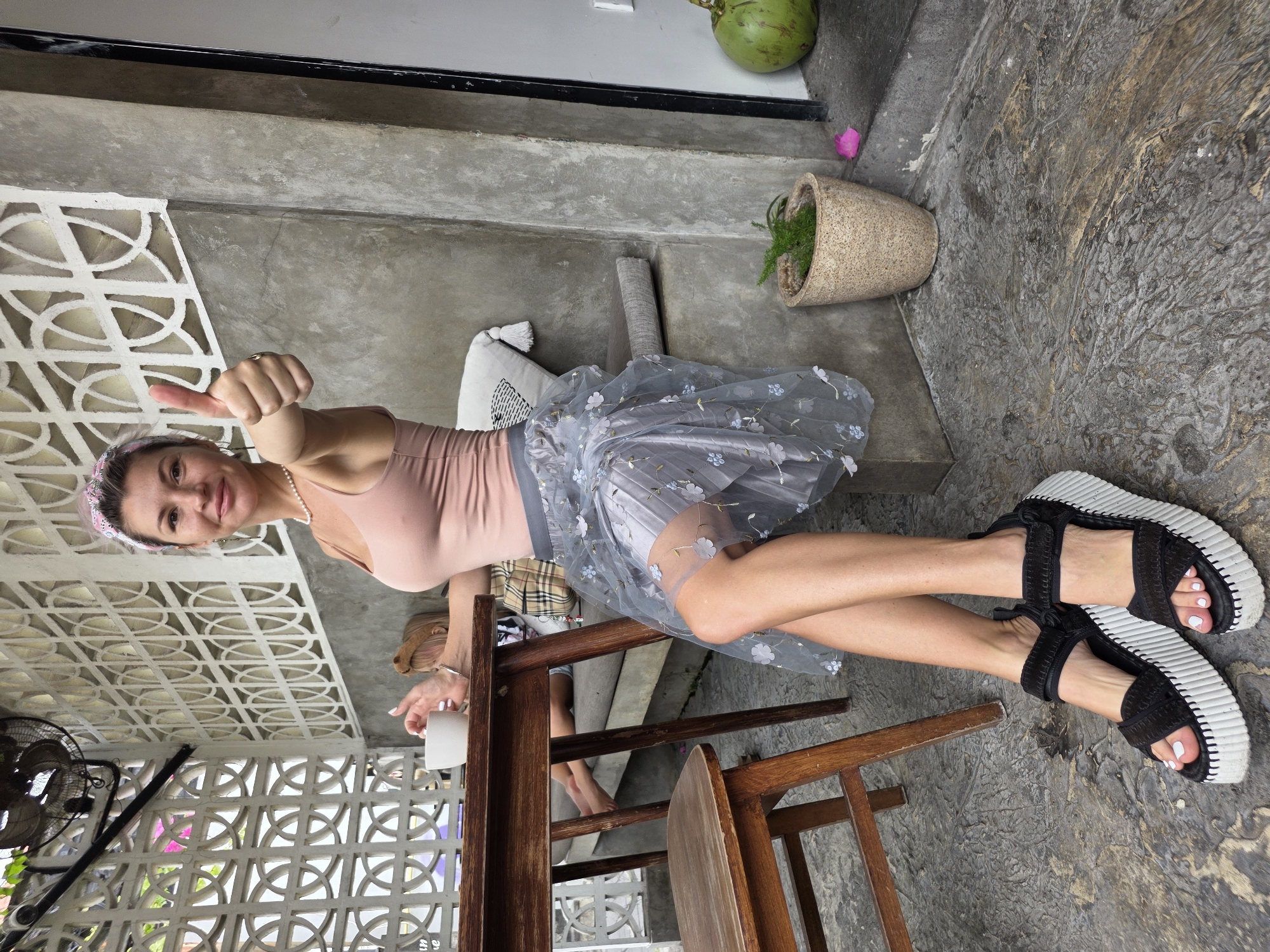A woman with pink-tinted hair, wearing a pink tank top and a grey, sheer, floral-patterned skirt, sits at a wooden table and gives a thumbs-up gesture to the camera. She wears black, platform sandals and a pearl necklace. In the background, a person with blonde hair sits on a bench behind the table, and a large green coconut sits on the ground near a doorway. The setting appears to be an outdoor seating area, possibly a cafe, with concrete walls and decorative latticework. The floor is a textured grey material. The location is identified as North Kuta, Indonesia.