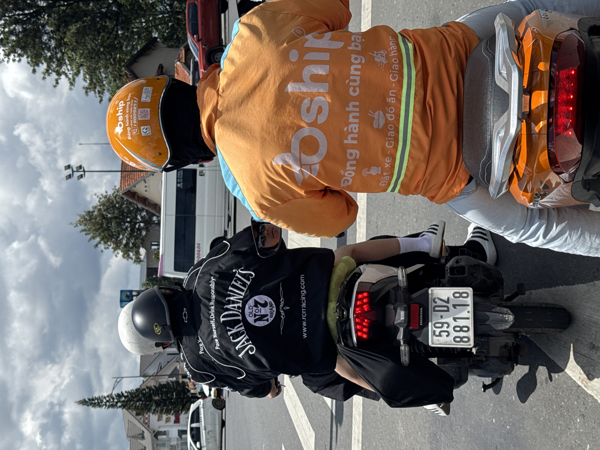 Two individuals are riding on a motorcycle in Da Lat, Vietnam. The person in front is wearing an orange jacket with "Gojek" logo and an orange helmet. The person behind is wearing a black jacket with "Jack Daniels" logo and a white helmet. The motorcycle has a license plate that reads "59-D2 881.18". The background shows buildings, trees, and cars, indicating an urban setting. The scene appears to be at a street intersection with traffic lights visible. The weather is cloudy.