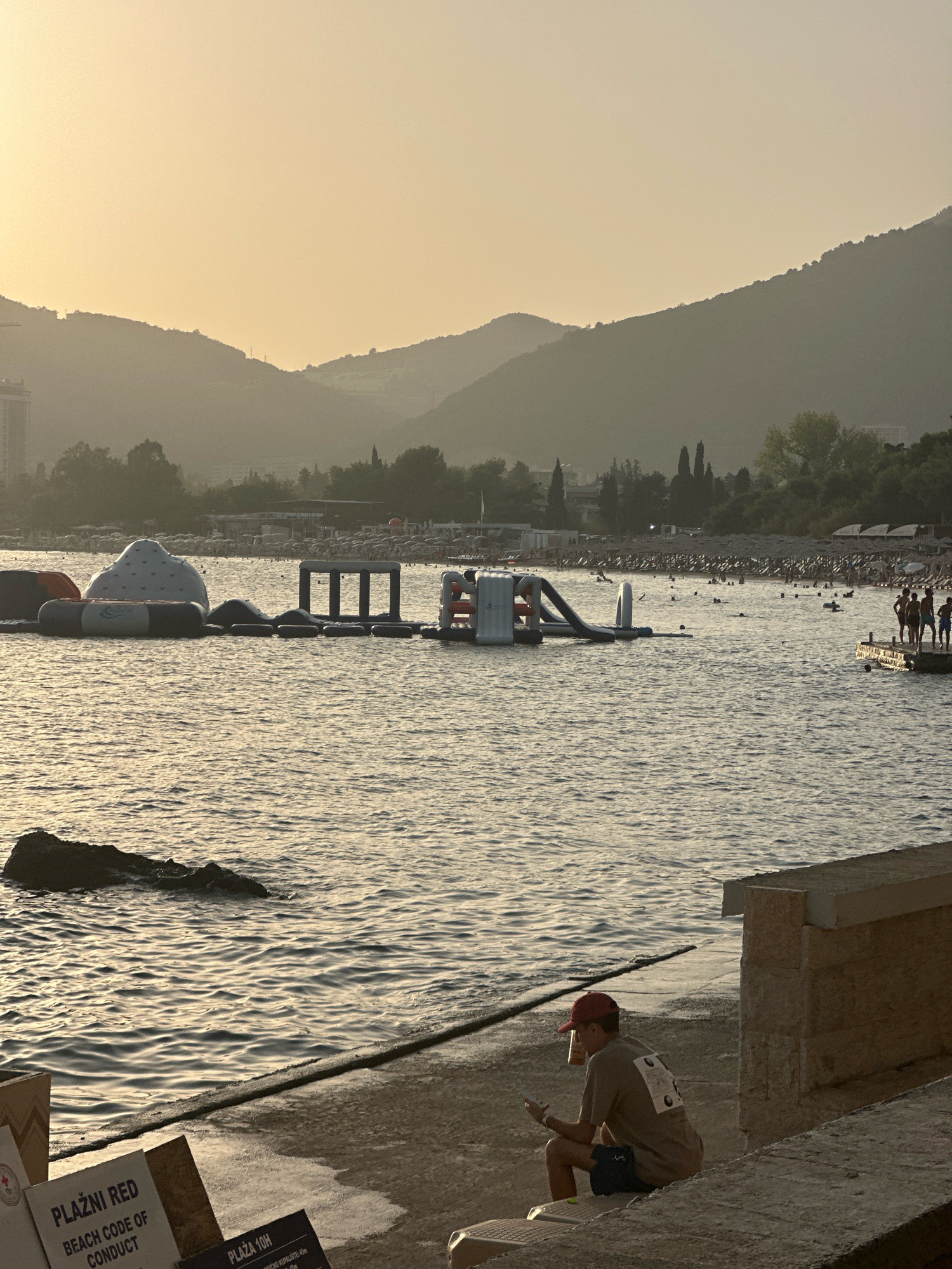 The image depicts a serene beach scene in Budva, Montenegro during what appears to be sunset. In the foreground, a young person wearing a red cap and a light brown t-shirt sits on a concrete ledge, looking at their phone. Behind them, the gentle waves of the Adriatic Sea create a shimmering surface.
Further out in the water, an inflatable water park with various structures like slides and climbing walls is visible. Several people are enjoying the water, swimming and interacting with the inflatable attractions. In the background, a beach lined with rows of sun loungers and umbrellas stretches along the coastline, suggesting a popular tourist destination. The scene is framed by rolling hills and mountains that are silhouetted against a hazy, golden sky, indicative of the setting sun. A few trees and buildings are scattered along the shore and the foothills of the mountains. The overall atmosphere is peaceful and relaxed, capturing a typical summer evening at the Montenegrin coast.