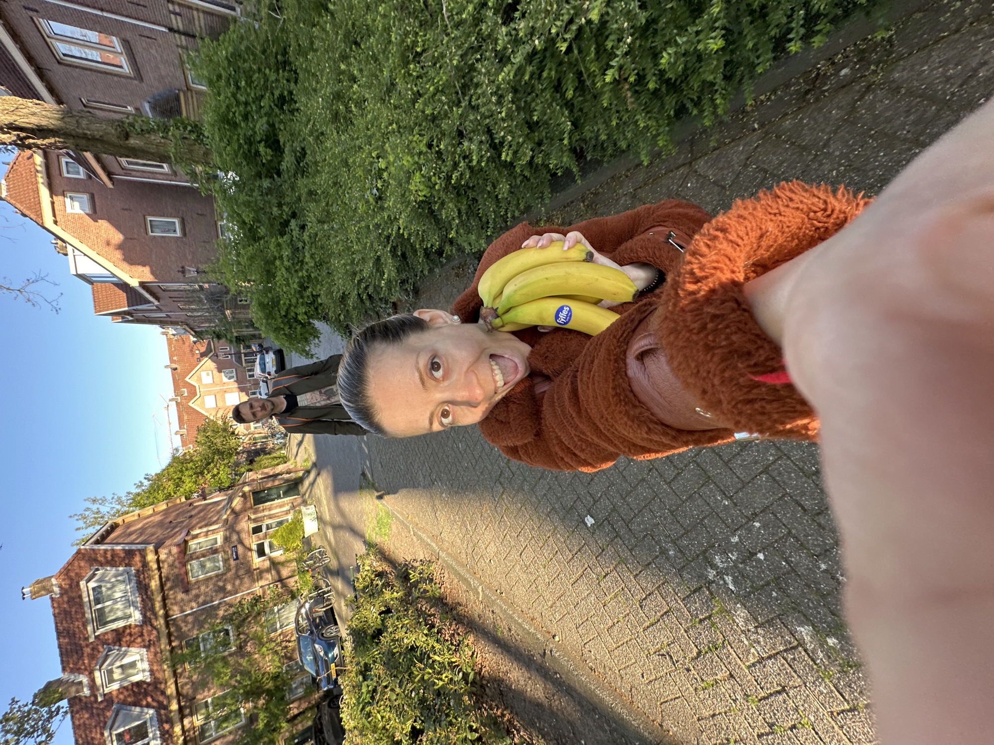 The image is a brightly lit, vertical selfie taken by a woman on a residential street, likely in Amsterdam, Netherlands. The primary subject is the woman in the foreground, holding a bunch of five ripe yellow bananas close to her face. She has her hair pulled back and is wearing a textured, reddish-brown fleece or faux fur jacket. Her expression is animated and playful, with a wide, open-mouthed smile revealing her teeth, and her eyes looking directly at the camera. A blue sticker with the brand name "Fyffes" is clearly visible on one of the bananas.
In the mid-ground, a second person, a man in a dark jacket, is walking away from the camera on the paved sidewalk. The background features a street lined with multi-story brick buildings, consistent with Dutch architecture, characterized by their distinct windows and roofs. Green trees and shrubs are abundant along the street. The time of day appears to be late afternoon or early evening, indicated by the warm, directional sunlight and the long shadows stretching across the patterned brick pavement. The sky is clear and blue. Several bicycles are parked along the curb, and a blue car is partially visible, adding to the urban residential setting. The overall scene conveys a cheerful and lively moment.