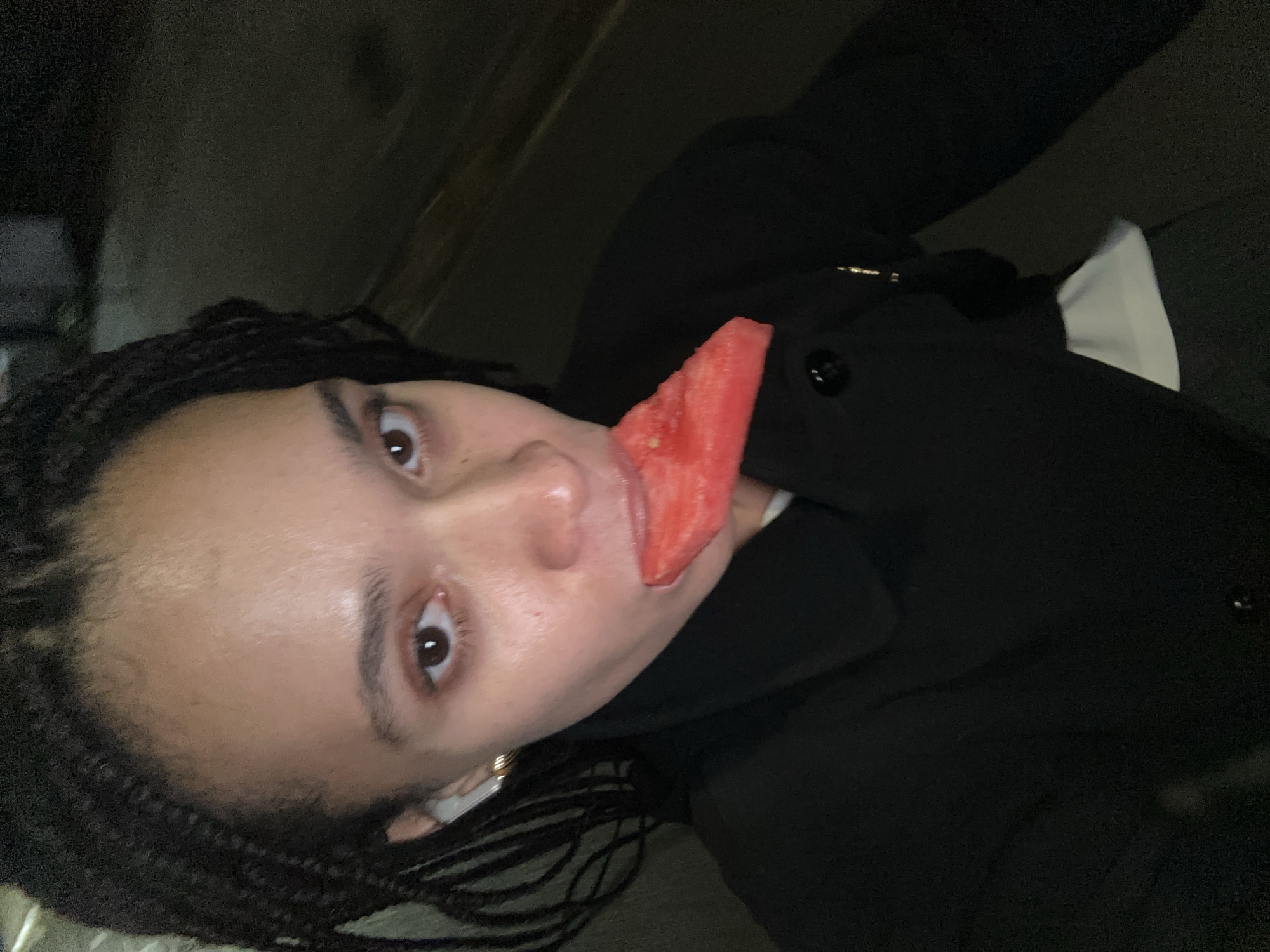 A close-up selfie shows a person with braided hair and brown eyes holding a slice of watermelon to their mouth. The person is wearing a black jacket and a white shirt is visible underneath. The background is dark and out of focus. There is no indication that the image was taken in London, United Kingdom.