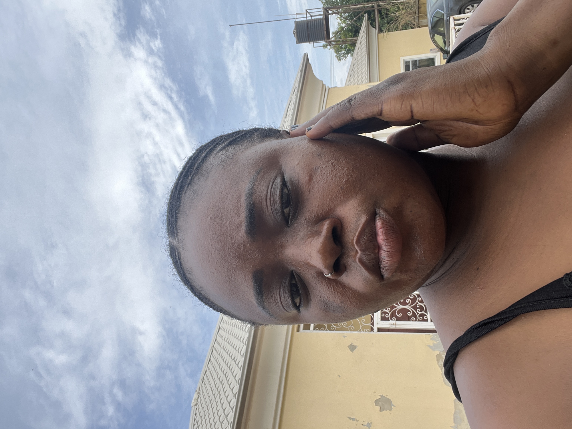 A portrait of a dark-skinned woman with short braided hair. She is wearing a black top and a nose ring. Her right hand is raised to her face, obscuring part of her forehead. The background includes a light yellow building, a water tank, a car, and a partly cloudy sky. The location appears to be in Abuja, Nigeria, based on the architectural style and surroundings.