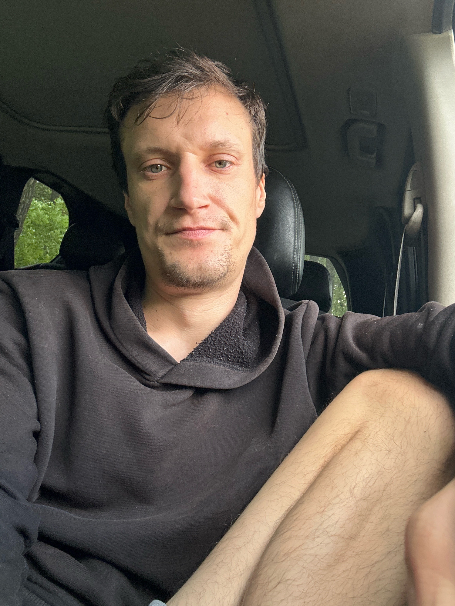 A man is sitting in the driver's seat of a vehicle, facing forward and looking at the camera. He has short, dark brown hair and light skin. He is wearing a black hooded sweatshirt and his right leg is visible, covered in light brown hair. The interior of the vehicle is dark, with a sunroof visible above. Through the windows, lush green trees can be seen, suggesting an outdoor, possibly rural, environment. The scene appears to be a candid selfie taken within the vehicle. The provided crops offer closer views of the man's face, the interior of the car, and his clothing and leg. There is no specific information in the image that places the location in Makiv, Ukraine.