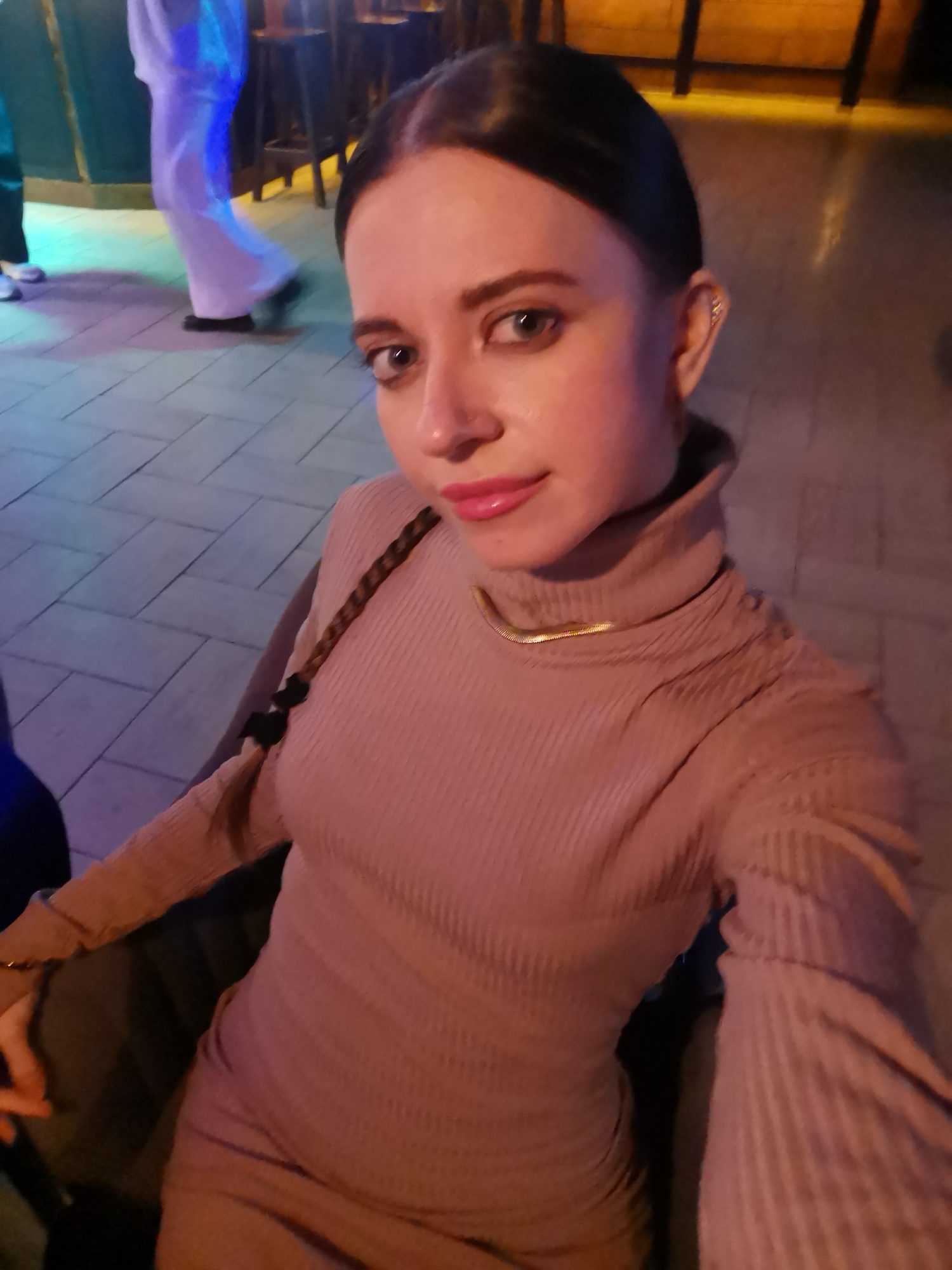 A woman is captured in what appears to be a selfie, looking directly at the camera with a neutral to slightly smiling expression. She has dark hair styled neatly, pulled back, with a visible braid resting over her right shoulder. She wears a light brown or nude-colored ribbed turtleneck dress or top, accessorized with a slender gold snake chain necklace and a small, intricate piercing on her upper left ear. Her lips are adorned with pink lipstick.
The setting appears to be an indoor or covered outdoor space, likely an entertainment venue such as a bar or club, given the dim and colorful artificial lighting. The floor is paved or tiled with a visible pattern, reflecting hues of blue, purple, and yellow from unseen light sources. In the background to the left, another person is partially visible, dressed in a long white garment, appearing to be walking. Hints of what could be bar stools or furniture are visible in the upper right background. The overall ambiance suggests an evening or nighttime event. No discernible text is visible in the image.