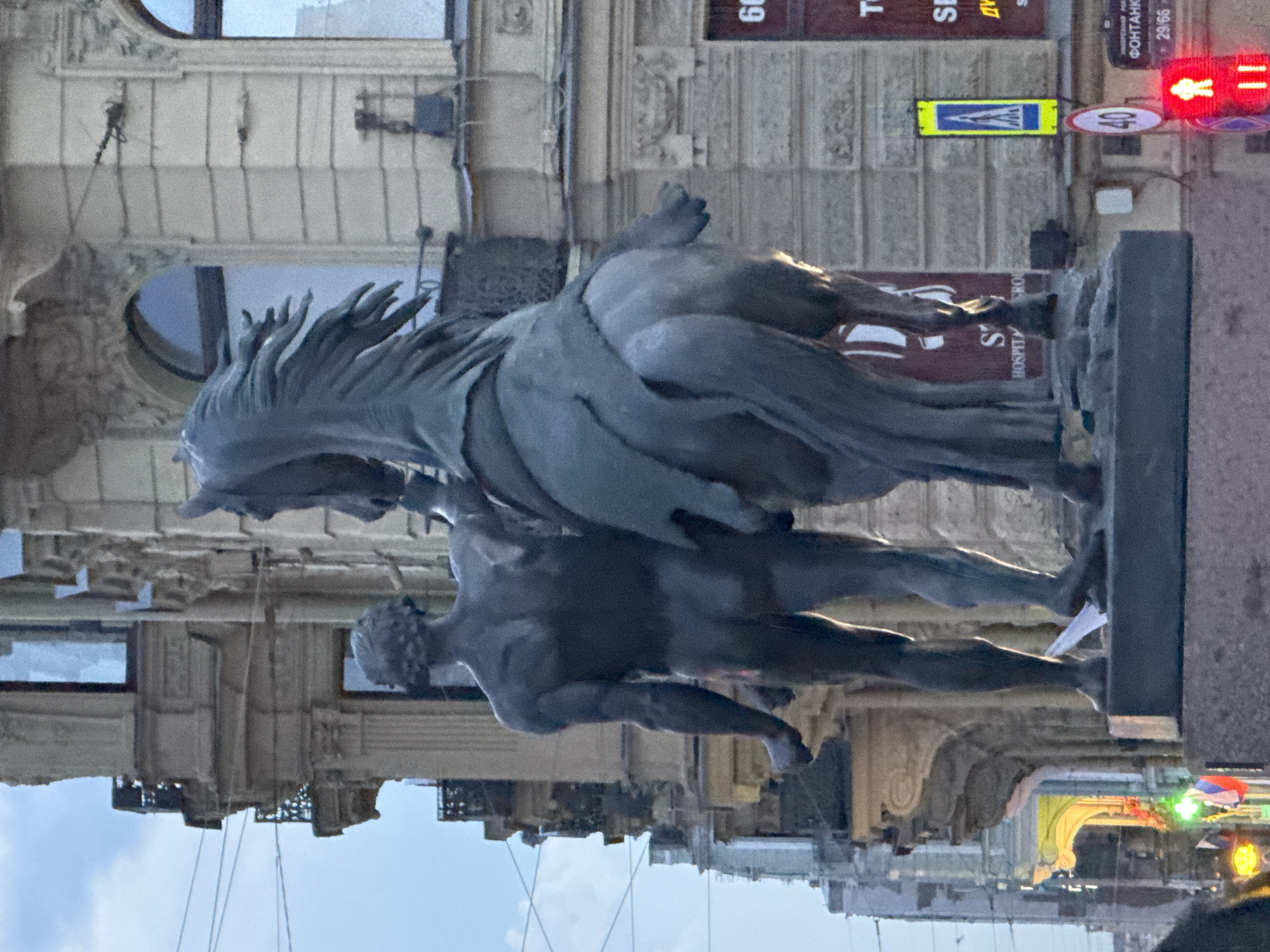The image captures a bronze equestrian statue in Saint Petersburg, Russia. The statue depicts a man standing behind a horse, with his left arm resting on the horse's back and his right arm extended downwards, holding the horse's reins. The man is nude, showcasing muscular definition. The horse is depicted in motion, with its mane flowing and its head turned to the left. The statue is positioned on a stone pedestal in front of a building with classical architectural elements, including columns, ornate carvings, and arched windows. The sky above is overcast. In the background, there are signs, including one with the number "60", a pedestrian crossing sign, and a traffic light displaying a red walking figure. The overall scene suggests a public square or street in the city.