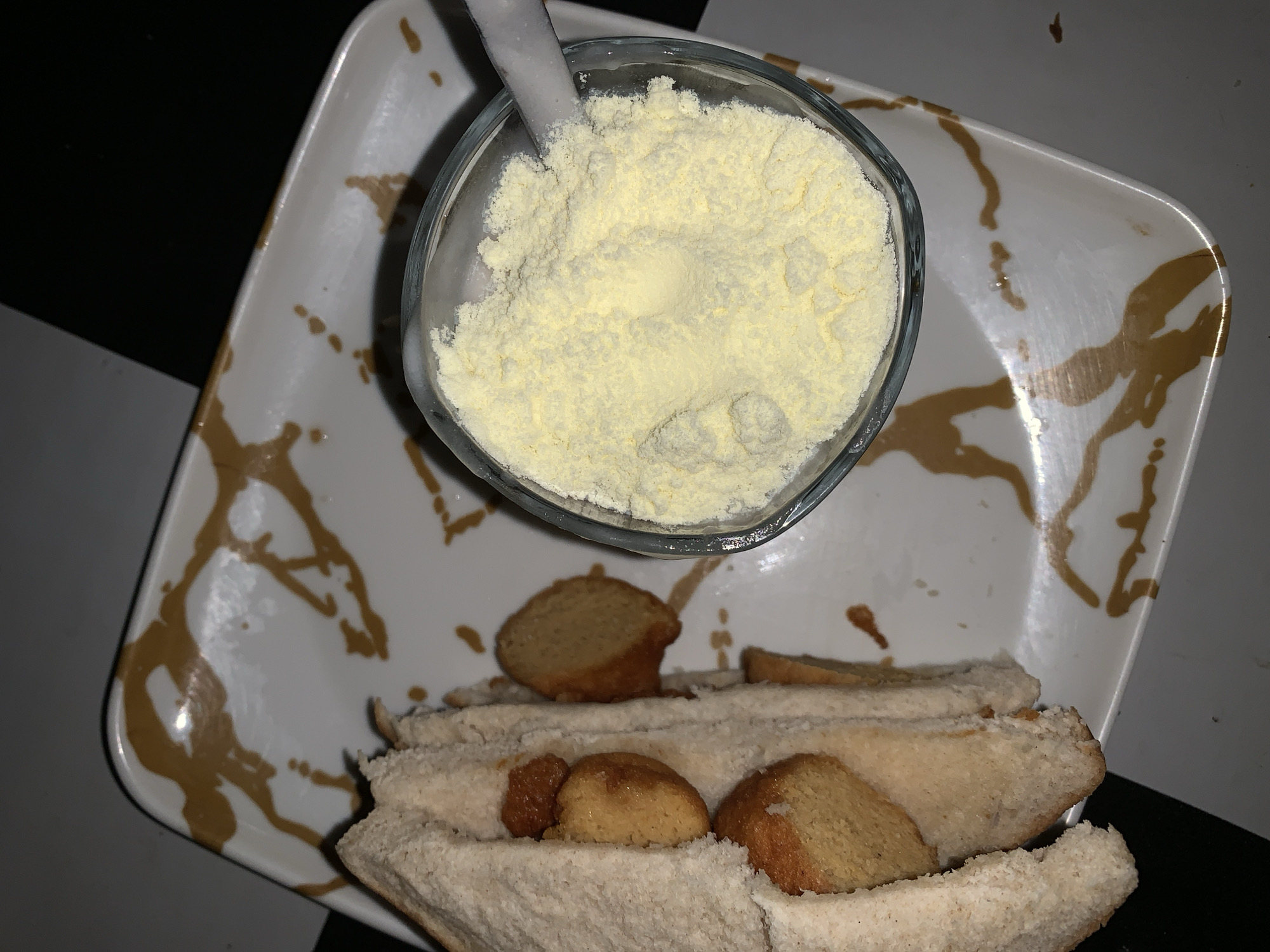 The image shows a glass filled with yellow powder, possibly powdered milk or a similar substance. A white spoon or utensil is partially submerged in the powder. Below the glass is a sandwich-like arrangement of bread slices containing round, brownish objects, possibly cookies or bread rolls. The food is arranged on a white plate with gold-colored abstract patterns. The background appears to be a mix of light and dark surfaces, indicating a room or a counter setting. There is no specific visual information to confirm the location in Rafin Par, Nigeria.