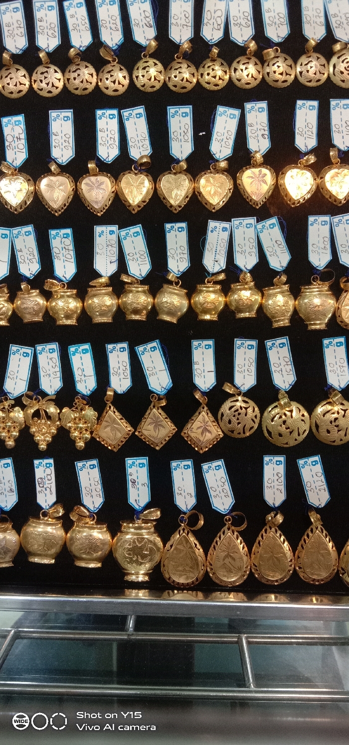 The image displays a static arrangement of approximately 60-70 yellow gold pendants, organized in six horizontal rows against a dark, non-reflective background, within a retail establishment in Maron, Indonesia. The pendants exhibit various designs, including spherical with filigree patterns, heart-shaped with engraved motifs (e.g., palm trees), vase-like forms, grape clusters, rhombus-shaped, and teardrop designs. Each pendant is accompanied by a small, rectangular white paper tag, attached with blue string, displaying handwritten notations such as "30 B %" and numerical values followed by "g," presumably indicating gold content and weight or price. The lower portion of the image shows the brushed metal frame and glass surface of a display cabinet. No individuals are present in the scene.