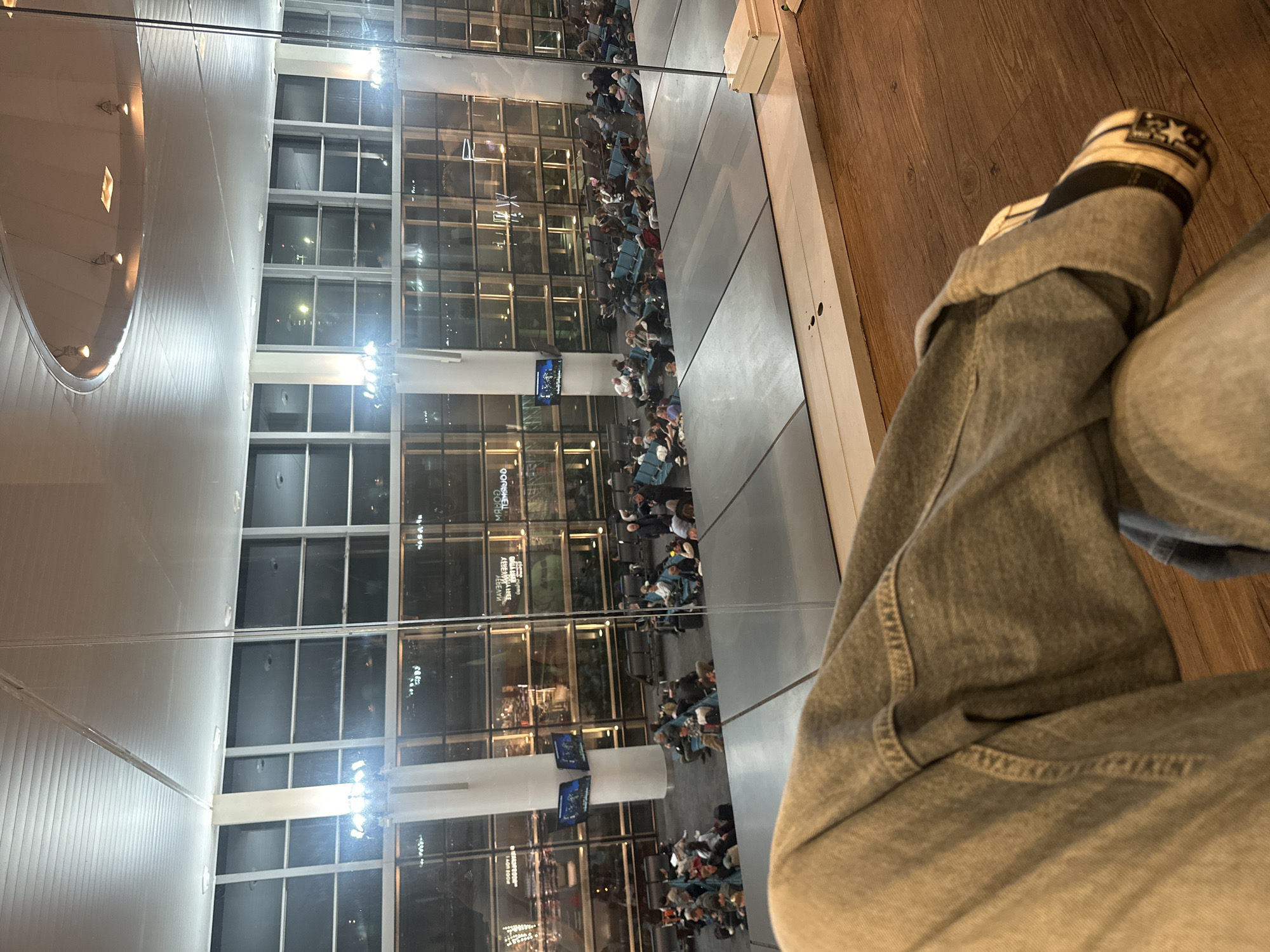 The image shows a person's legs and shoe in the foreground, with a mirrored reflection of an airport terminal in Yerevan, Armenia. The terminal has large windows showing waiting areas with seated passengers. There are televisions mounted on columns. The reflection shows interior architectural details. The setting appears to be inside a building with wood flooring.