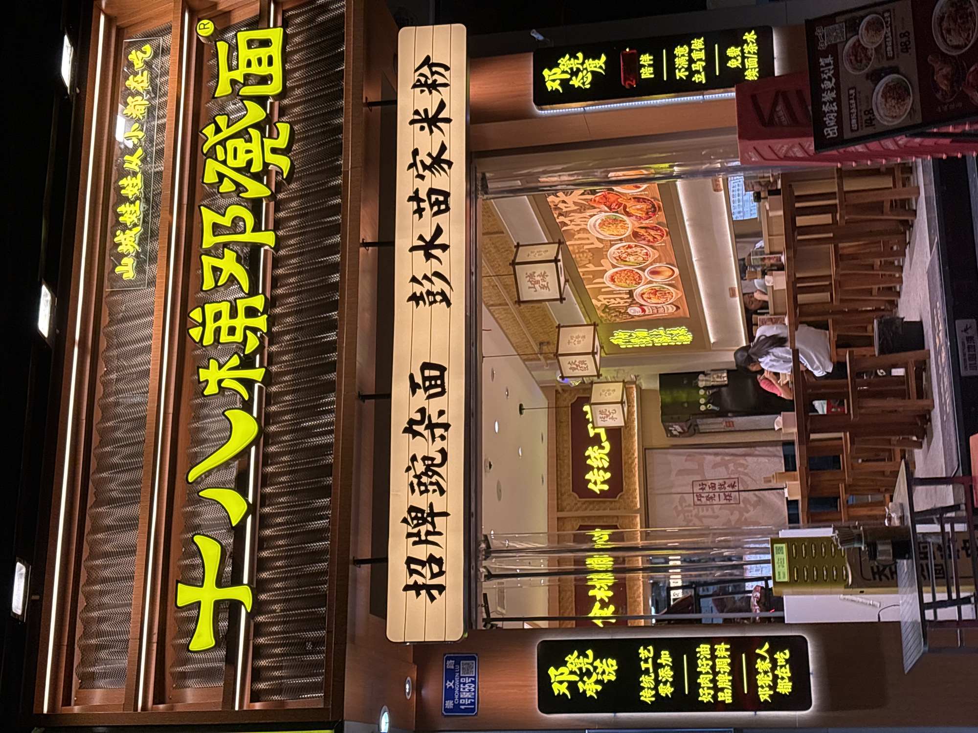 The image shows the exterior of a restaurant in Nan'an District, China, at night. Two lit signs on the left read "十八梯邓凳面" and "招牌豌杂面 彭水苗家米粉". A smaller sign below has the street address and name "崇文路" and "CHONGWEN LU". Below the right sign is another smaller sign with phrases about the restaurant. Through the glass front, people are sitting and eating inside, with wooden tables and chairs visible. Illuminated menu boards display dishes above the diners. Several hanging decorative lamps are hanging inside.