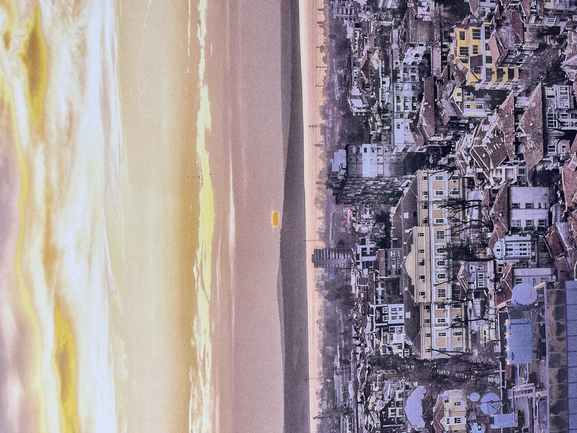 The image depicts an aerial view of Burgas, Bulgaria, situated along the coastline. The city's buildings, varying in size and structure, are densely packed together with many having reddish-brown roofs. To the left of the city is a body of water, likely the Black Sea, with a hazy, golden sky above it.