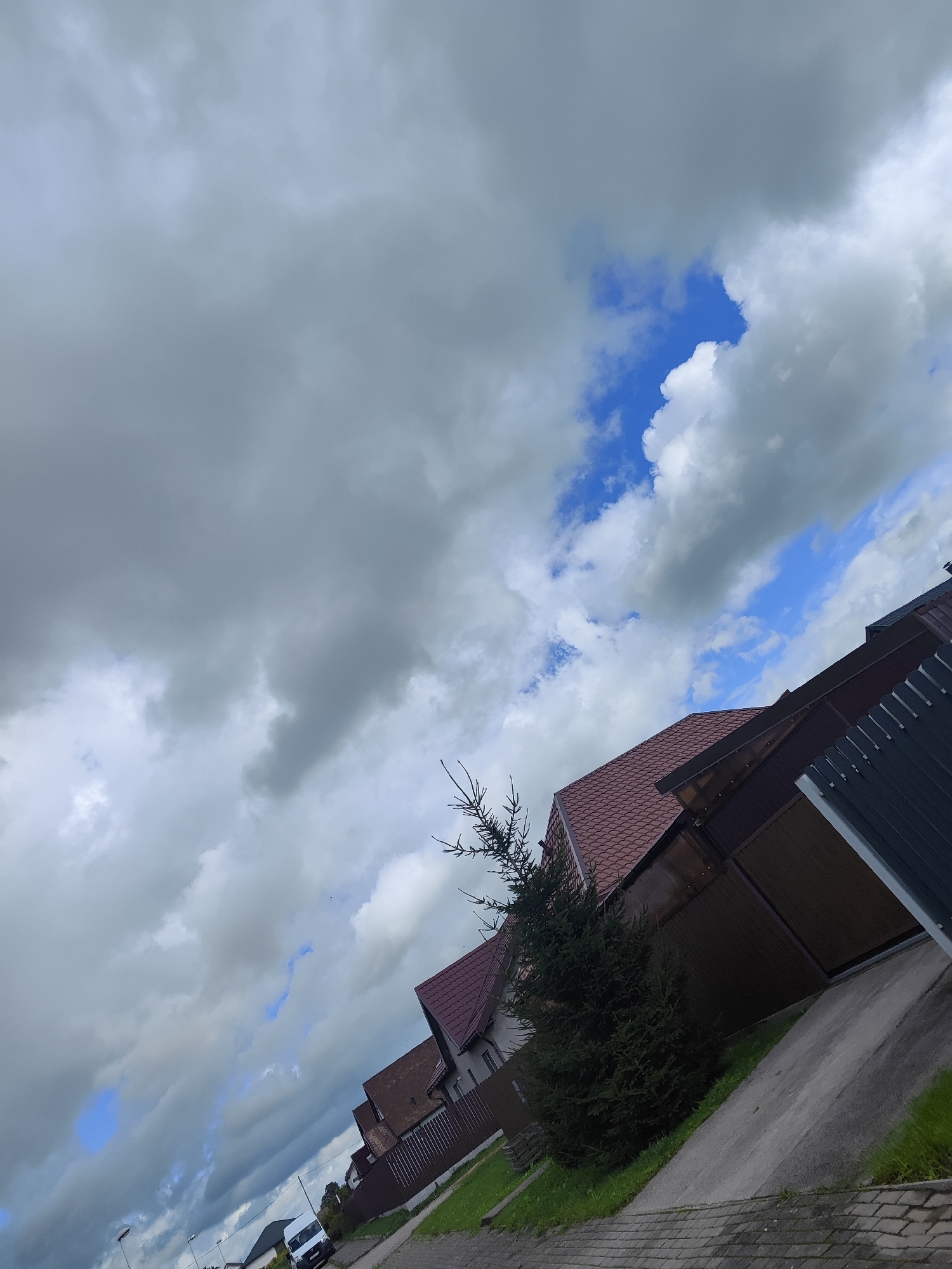This is a photograph taken from a street in Gvardeysk, Russia, during a cloudy day with patches of blue sky. The image is tilted, giving it a dynamic perspective. In the foreground, a brick-paved walkway runs along a sloped asphalt driveway. To the right of the driveway, there is a dark metal fence. Beyond the fence and driveway, several houses are visible, characterized by their reddish-brown tiled roofs and brown wooden fencing. A dark green coniferous tree stands near one of the houses. In the distance, a white van is parked on the street. The sky is dominated by heavy, gray clouds, but there are breaks that reveal patches of bright blue sky.