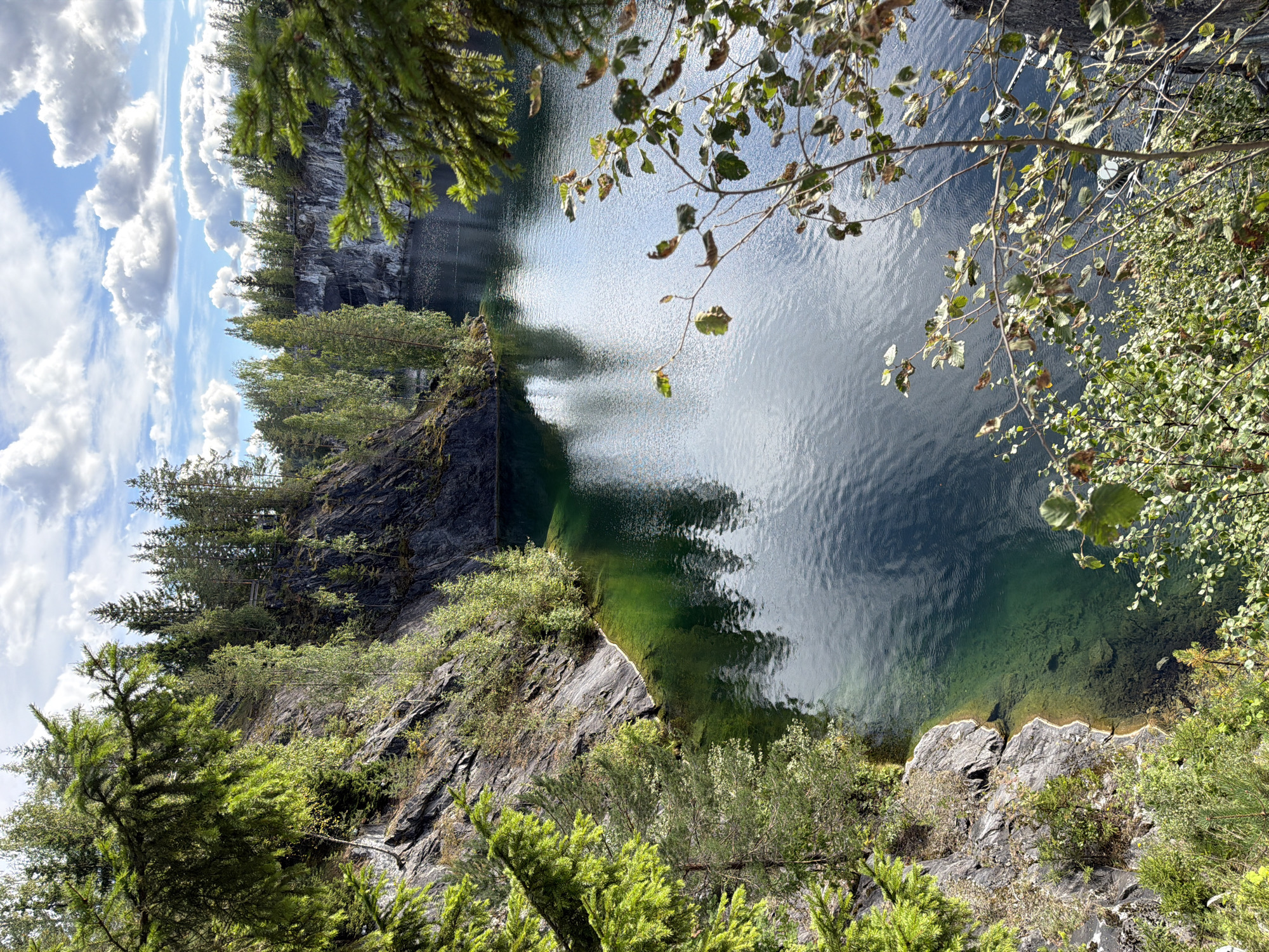 The image depicts a serene natural landscape at the Ruskeala Mountain Park, likely in the Republic of Karelia, Russia. The scene is dominated by a body of calm, emerald-green water, reflecting the surrounding environment. Steep, rocky cliffs rise from the water's edge, sparsely covered with lush green vegetation and a variety of trees, including coniferous and deciduous species. The sky above is a vibrant blue, dotted with fluffy white clouds, suggesting a pleasant, sunny day. The foreground is framed by the branches of trees, some with vibrant green needles and others with scattered dry leaves, indicating it may be autumn. The overall impression is one of natural beauty and tranquility, with the contrasting textures of rock, water, and foliage creating a visually appealing composition. There are no people or visible man-made structures apart from a hint of what might be a viewing platform or bridge in the distance along the cliff face.