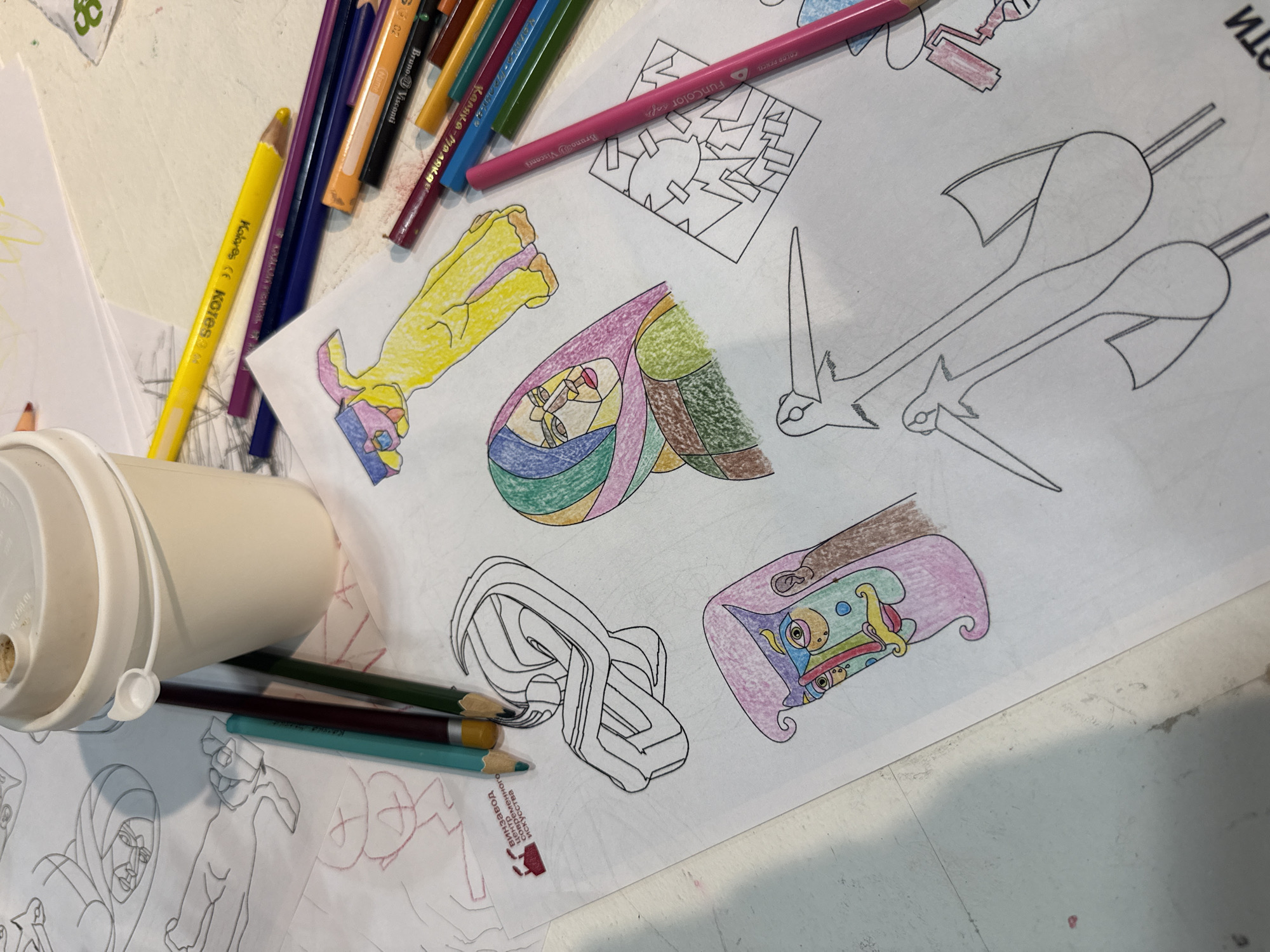 The image depicts a close-up view of a table with various coloring pages and colored pencils, suggesting a creative activity, possibly at home or a creative space. A disposable coffee cup is prominently placed on the upper left, with its lid slightly ajar. Several coloring pages are spread out, showcasing unfinished drawings of abstract figures, a stylized face with geometric patterns, and a pair of birds with long necks. Colored pencils are scattered around, with some actively used to color parts of the drawings. One coloring page features a logo with Russian text that reads "Винзавод Центр Современного Искусства," which translates to "Winzavod Center for Contemporary Art." This logo indicates that the coloring pages likely originate from or are associated with the Winzavod Center for Contemporary Art in Moscow, Russia. The scene is brightly lit, and the focus is on the art supplies and the creative process.