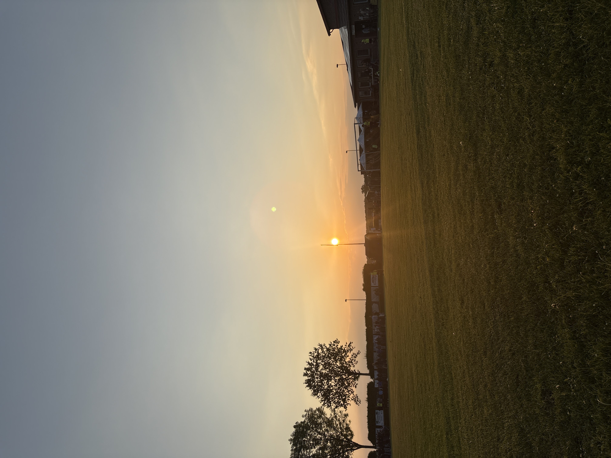 The image shows a sunset over a grassy field in Wegberg, Germany. The sun is low on the horizon, casting a warm glow over the scene. In the foreground are two trees. In the background is a building.