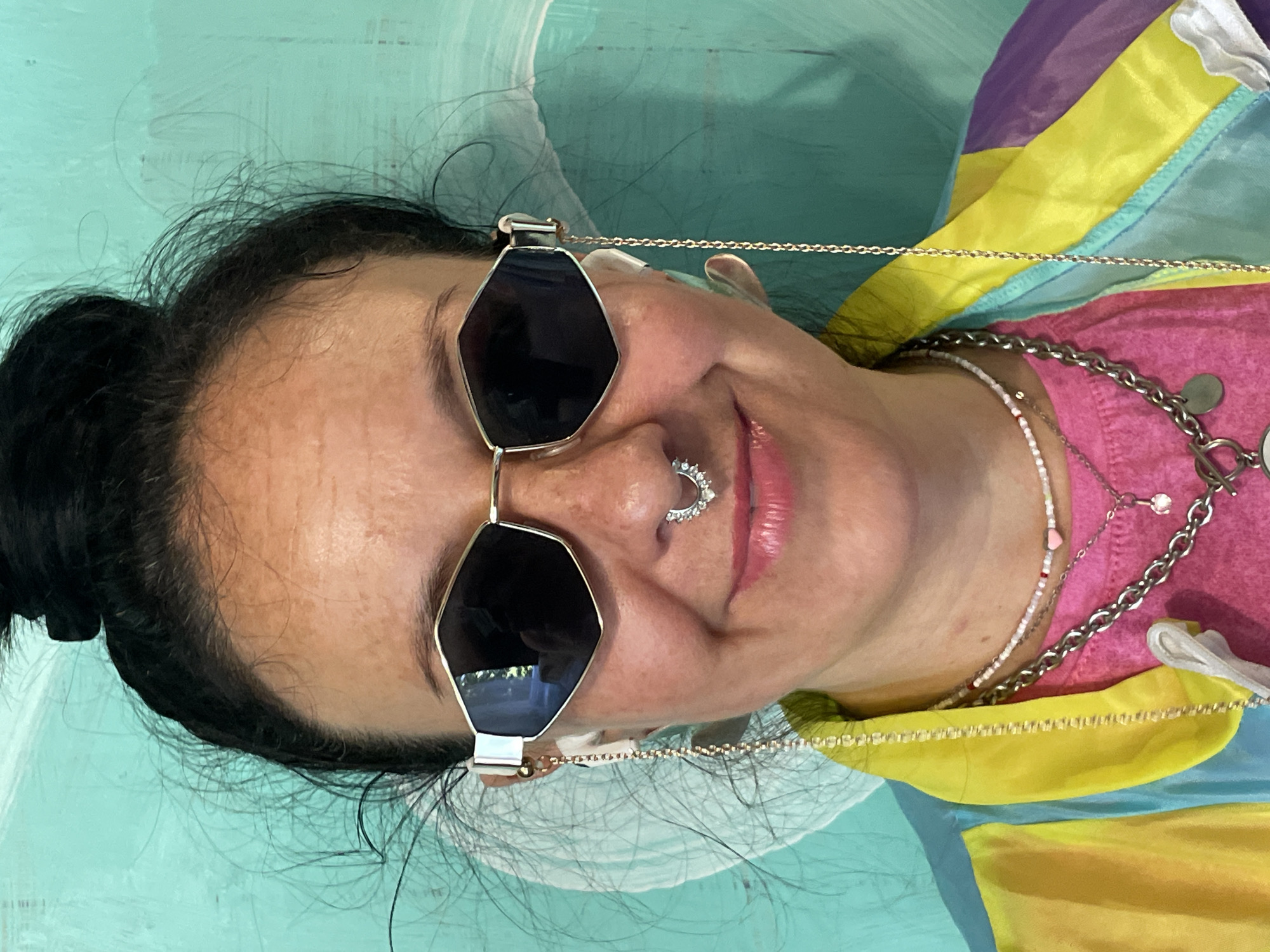 A medium close-up, head-and-shoulders shot captures a woman with dark hair pulled into a bun. She wears large, dark-lensed sunglasses with a gold hexagonal frame. A silver septum ring with small rhinestones adorns her nose. She has on a bright pink t-shirt, over which a vibrant jacket with sections of yellow, blue, and purple is visible. Several necklaces are layered around her neck, including a delicate beaded one with a pink heart pendant and a thicker silver chain. Chains also hang from her sunglasses, attached to what appear to be wireless earbuds. The background is a textured, teal-colored wall with abstract white brushstrokes. The lighting suggests an indoor setting, possibly a studio. There is no discernible indication of this location being in Saint Petersburg, Russia.