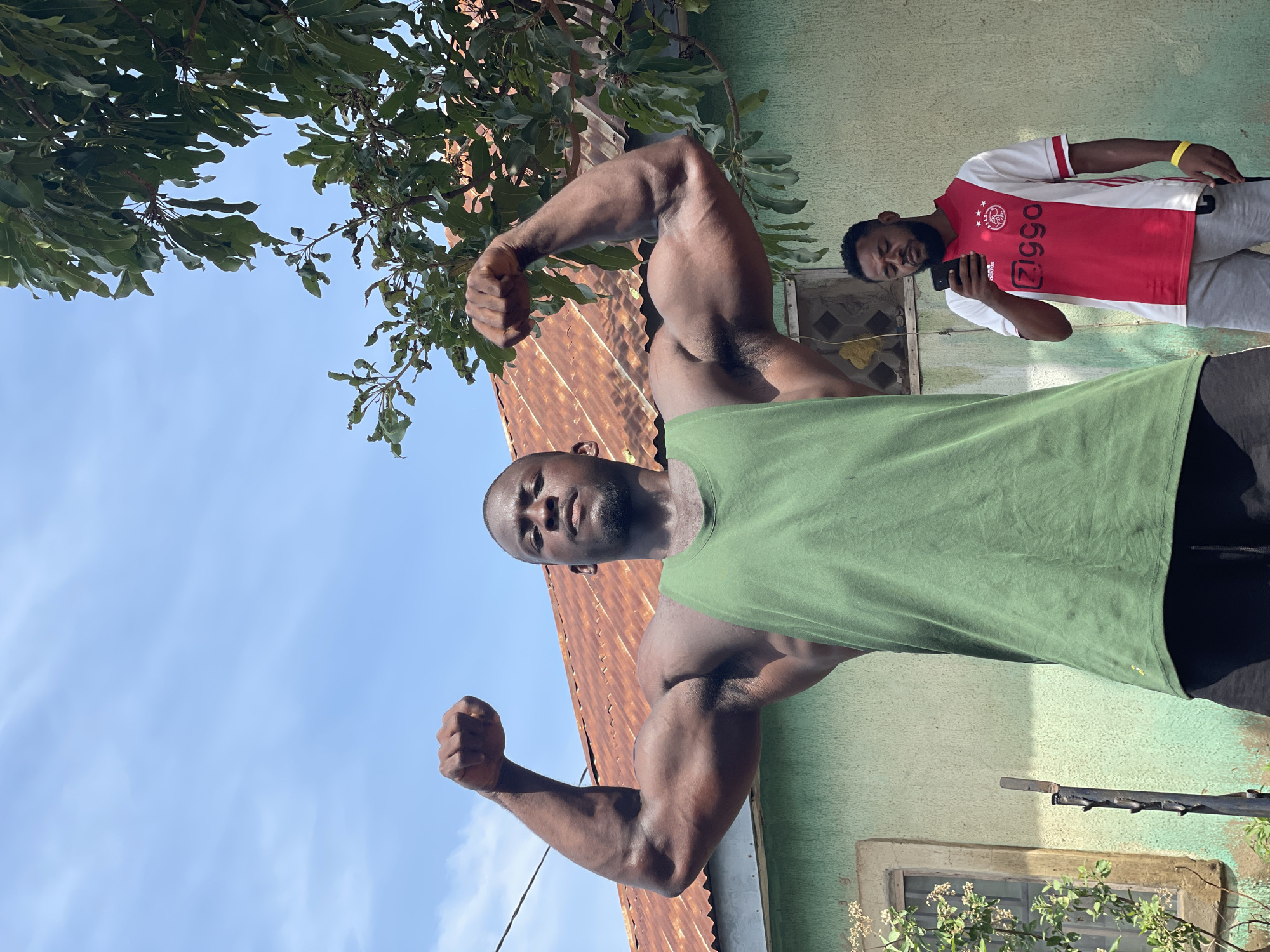 In Akwanga, Nigeria, a man in a green tank top flexes both arms, displaying large biceps. A second man, wearing a red and white Ajax soccer jersey, looks at a smartphone behind the first. Both men are in front of a building with a rusty metal roof and painted green walls. Foliage is visible in the top left corner, and a blue sky is visible.