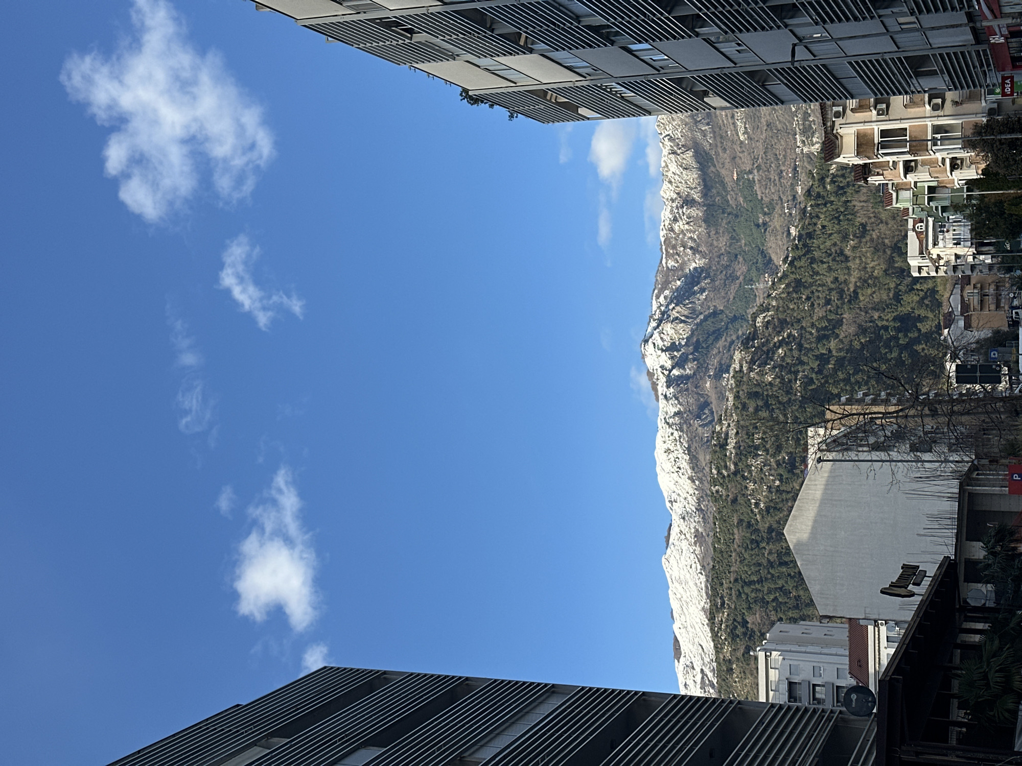 The image captures a view in Budva, Montenegro, characterized by a juxtaposition of urban architecture and natural landscape. The left side of the frame is dominated by a bright blue sky dotted with a few white clouds. The bottom and top corners are framed by modern buildings with dark, horizontal slats along their facades, giving a sense of urban confinement. On the right, a mountain range covered with patches of snow and dense green vegetation rises dramatically. The base of the mountain merges with the urban development of Budva, where residential buildings and trees are visible. The overall impression is a dynamic contrast between the modern urban environment and the rugged natural beauty of Montenegro.