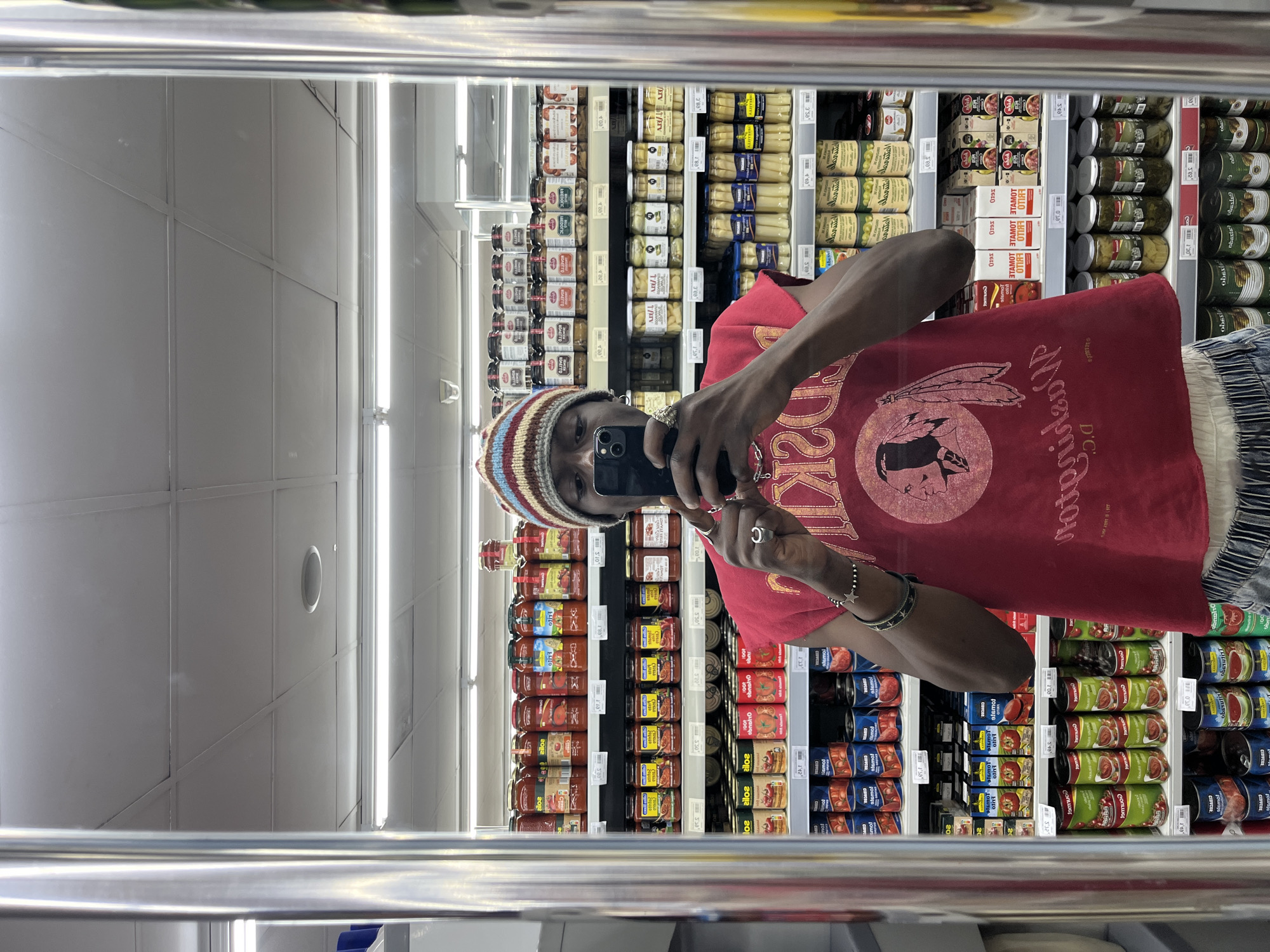 The image displays a mirror selfie taken by a person in a supermarket or grocery store aisle. The photograph is oriented vertically, with the top of the image being the right side of the scene.
The main subject is a person, likely a young adult with dark skin, whose face is partially obscured by the smartphone they are holding to take the picture. They are wearing a multi-colored striped beanie (featuring brown, beige, blue, and red hues), a red t-shirt with a prominent graphic design, and light-colored, possibly striped or textured pants or shorts. The graphic on the t-shirt includes a stylized depiction of a Native American chief's head in a circular emblem, along with text that appears to be "DC" and other mirrored words like "Washin" (likely part of "Washington") and "CIDVCOR", which are part of the mirrored design. The person has several rings on their fingers and bracelets on their left wrist.
The setting is an indoor grocery store aisle, evident from the numerous shelves stocked with various jarred and canned goods, predominantly food items like sauces, pickled vegetables (e.g., asparagus, olives), and other preserves. The reflections in the mirror show rows of products, with prices visible on many shelf labels (e.g., "3.10", "4.00", "3.20", "2.90", "2.70", "3.30", "5.10", "4.50", "2.50"). Many product labels show mirrored text, such as "stamot" (which is "TOMATO" reversed), "CIDVCOR", "CONLIGE", "RAS", and "obsle" (likely a mirrored brand name). Fluorescent lighting fixtures are visible on the ceiling and reflecting on the store surfaces. The overall impression is casual and everyday, consistent with a shopping trip in Barcelona, Spain, given the product types and pricing format.