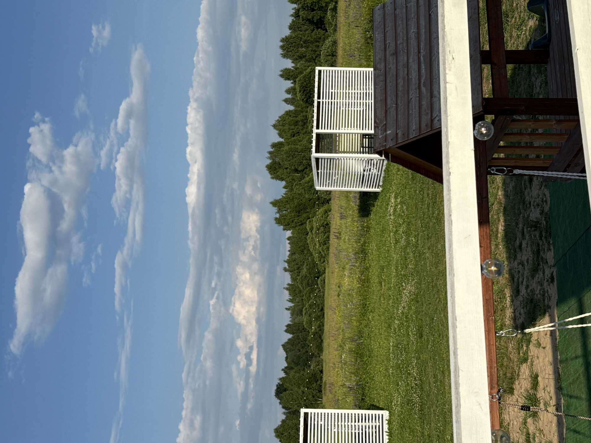 This is a vertical, eye-level, outdoor photograph taken in Колокша, Russia. The scene depicts a natural environment with some man-made structures. In the foreground, a wooden railing with decorative glass light bulbs spans the width of the frame. Below the railing, elements of a wooden play structure are visible, including ropes and a dark green swing seat.
The midground features a lush green lawn interspersed with patches of sandy soil and a synthetic green mat. Beyond the lawn, a field of grass with scattered purple flowers stretches towards a line of trees. A white, slatted modern structure, possibly a gazebo or screen, stands in the middle ground to the left. To its right, a similar white, slatted structure is visible, partially obscuring a dark wooden roof of another structure, which appears to be a playhouse. A string of white lights is visible running horizontally across the field of flowers.
The background is dominated by a dense forest of green trees under a blue sky with scattered white clouds. The lighting suggests it is late afternoon or early evening, with the sun casting shadows. There are no people or animals present in the image.