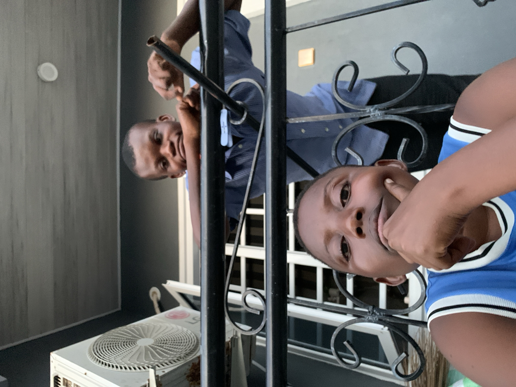 An image, originally rotated 90 degrees clockwise, captures two males, likely a father and son, posing playfully for the camera. They appear to be on a balcony or behind a railing of a building in Ibasa, Nigeria.
In the foreground, slightly to the right, a young boy with dark skin and short hair smiles broadly, looking directly at the viewer. He wears a blue t-shirt with white and black striped trim on the collar and sleeves. His left hand is raised towards his face, possibly covering his mouth or chin in a shy or thoughtful gesture.
Behind an ornate black metal railing, a man, also with dark skin and short hair, smiles and looks at the camera. He is wearing a light blue collared shirt, and his arm is visible, appearing to hold onto the railing. The decorative metal railing features several curved scrolls.
In the background to the left of the man, a plain dark gray wall is visible, with a small white circular object, possibly a light fixture or sensor, mounted high on it. In the lower left corner of the image (when oriented correctly), an outdoor air conditioning unit with a circular fan grate is visible, appearing slightly worn. White window or door frames can also be seen in the background, beyond the railing. The scene is well-lit, suggesting daytime. No discernible text is visible on any objects or clothing. Both individuals convey a sense of happiness and warmth.