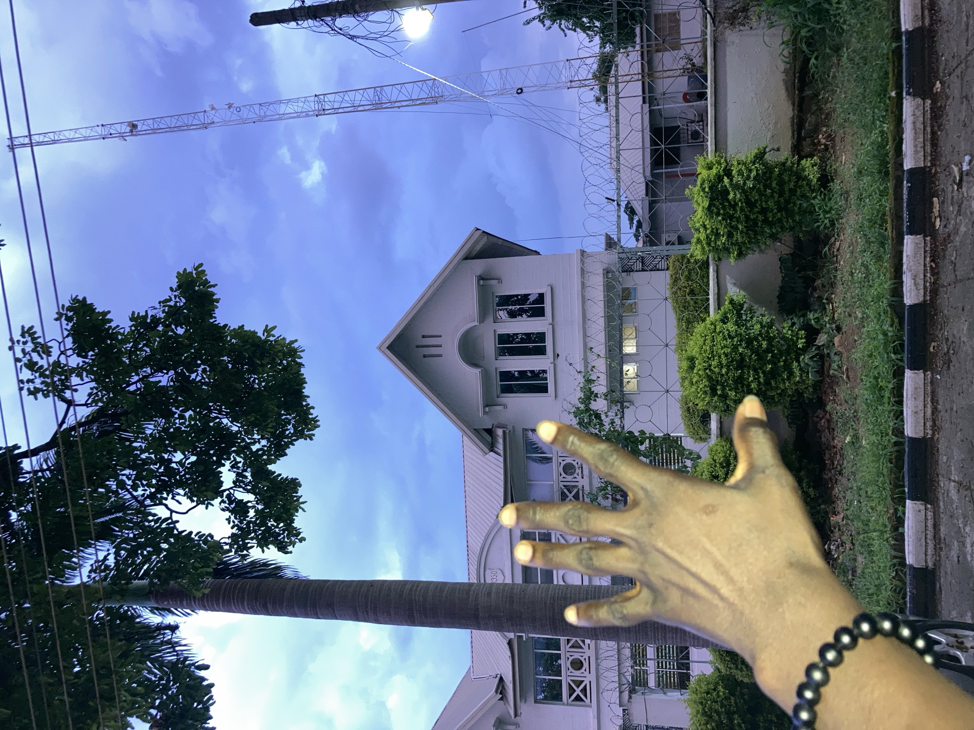 In a medium shot captured from a low angle in Jos, Nigeria, a person's outstretched left hand with a black beaded bracelet is in the foreground, obscuring part of the scene. The hand is reaching towards a white building with multiple windows and a distinctive triangular pediment. Behind the building, a tall, lattice radio tower stands against a cloudy, twilight sky. A palm tree trunk is visible on the left side of the frame, its leaves reaching towards the top. The ground in front of the building is lined with neatly trimmed green bushes, and a barbed-wire fence runs along the perimeter. The overall impression is a juxtaposition of man-made structures and natural elements in an urban setting.