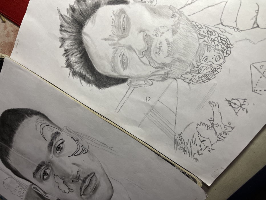 This image depicts two open sketchbooks displaying graphite portrait drawings, situated on a flat, reddish-brown surface in an indoor setting within the city of Jos, Nigeria.
The left page presents a graphite portrait of a male individual, shown from the chest up, with short, dark hair, prominent eyebrows, and an upward gaze. Distinct facial features include a small mustache and a septum piercing. Stylized, abstract markings are visible on the subject's left cheek and right temple. A faint, incomplete sketch of a human head is discernible in the upper left background.
The right page displays another graphite portrait of a male individual, characterized by spiky hair, squinted eyes, and a pursed mouth. The subject has a visible beard and mustache. Below the chin, the collar area is intricately detailed with a complex pattern incorporating multiple small faces and geometric elements. Adjacent to this portrait, incomplete sketches include a winged creature, a pyramidal symbol containing an eye, and other linear geometric forms.