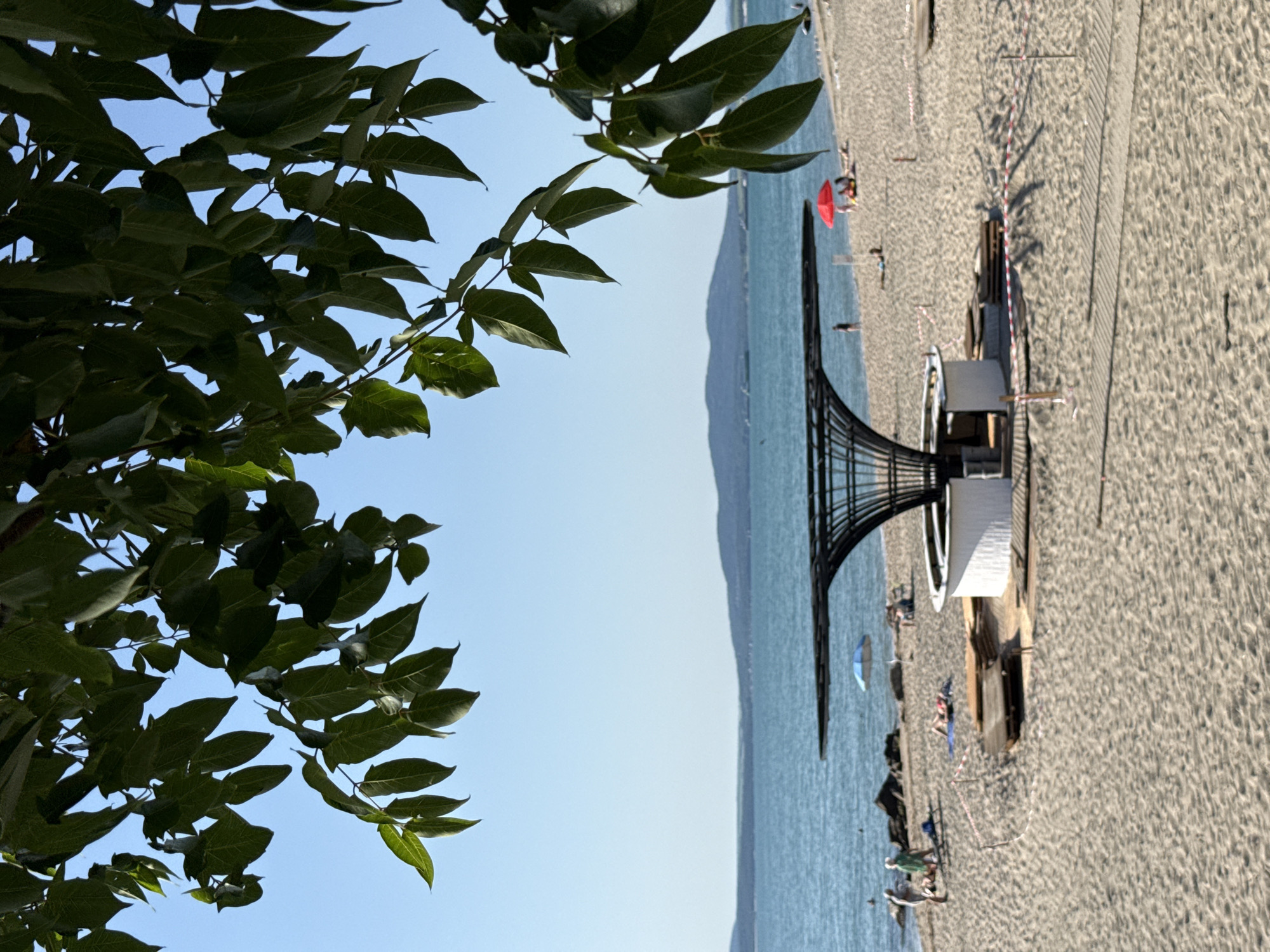 The image depicts a sunny day on the beach, with lush green tree branches partially obscuring the upper portion of the frame. The foreground is dominated by sand, with a pathway of wooden planks visible in the lower right. In the midground, a circular white structure with an abstract, black, umbrella-like roof stands prominently. Several people are scattered across the beach, some sunbathing, while others stand near the water's edge. A bright red umbrella and a blue and white umbrella are visible among the beachgoers. The background features a calm, blue sea stretching to the horizon, with a distant, tree-covered mountain range. The sky is clear and blue.
The scene is identifiable as the beach adjacent to the Sea Garden in Burgas, Bulgaria. The architectural style of the white structure with the unique roof is characteristic of some of the modern installations found along Burgas's coastline. The specific location is likely near the central part of the beach, given the presence of amenities and the overall arrangement of beach infrastructure.