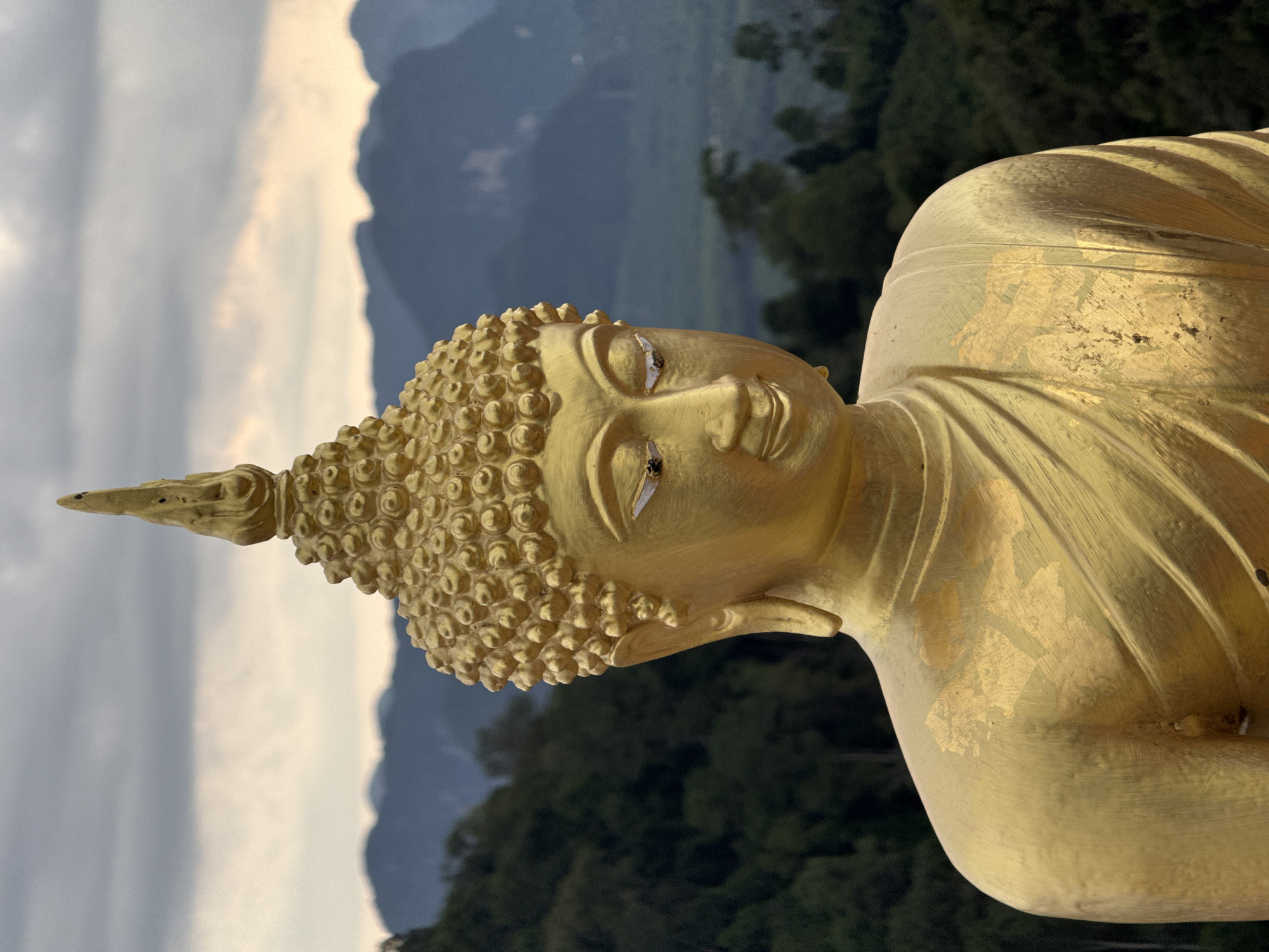 A close-up, eye-level shot shows the upper half of a large, gold-colored Buddha statue. The statue is positioned outdoors, with a backdrop of lush green trees and dramatic, hazy mountains under a cloudy sky. The statue's face is serene, with downcast eyes. The top of its head is adorned with intricate curls and a pointed finial. The gold surface of the statue shows some wear, with patches of gold leaf missing in places, particularly on the chest and shoulder area. The statue is located at a viewpoint in Ban Khao Tang, Thailand, likely at a temple or elevated scenic spot. The lighting suggests it is either early morning or late afternoon, with diffused light from the overcast sky.