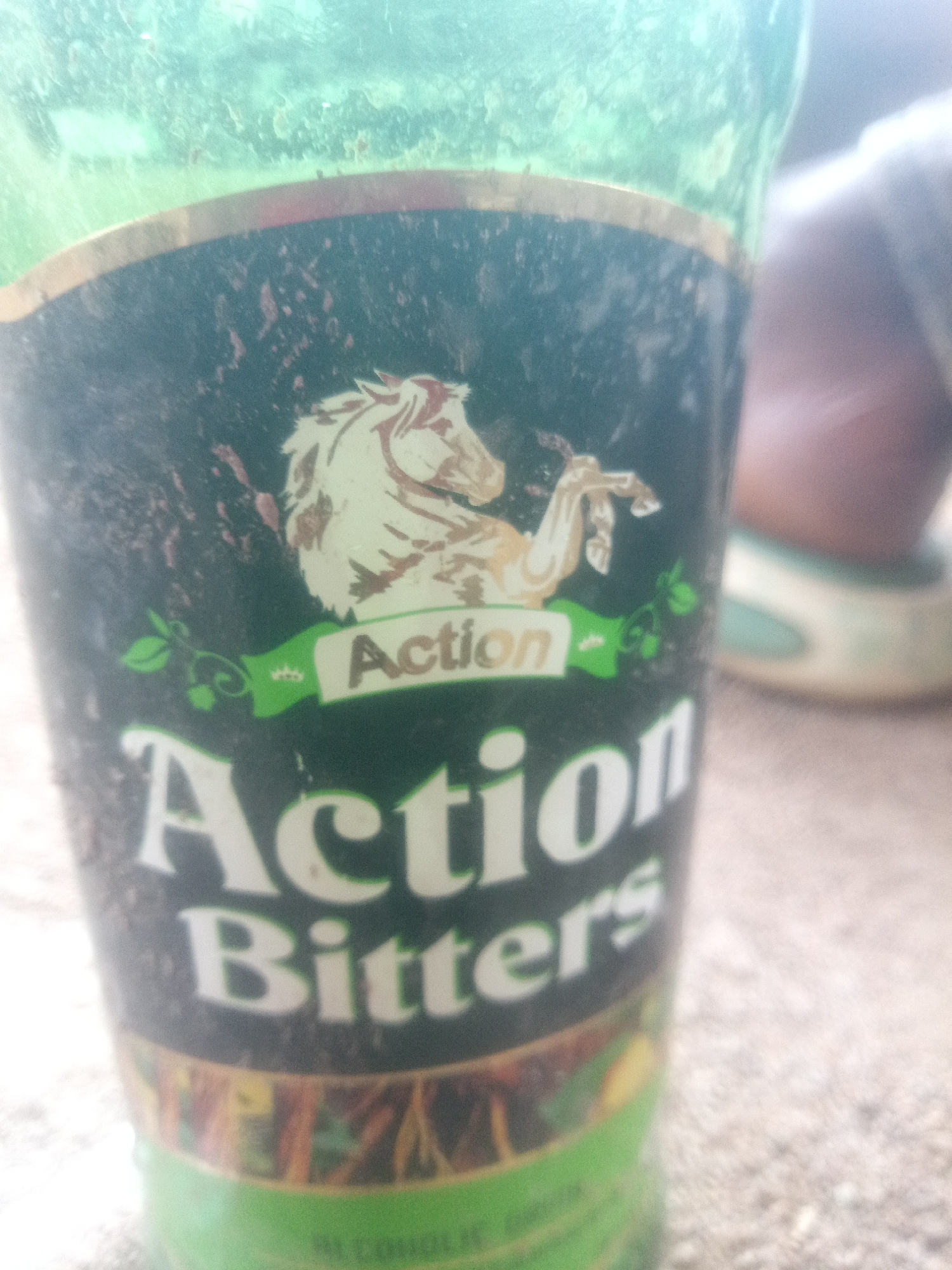 The image shows a close-up of a green bottle with a label that reads "Action Bitters." A stylized image of a horse is visible on the label, along with decorative elements and smaller text. The bottle appears to be dirty or smudged. The background is blurred, showing what looks like a surface of sand or gravel, and a person's leg and shoe. The lighting suggests an outdoor setting. The location is not discernible from the image.