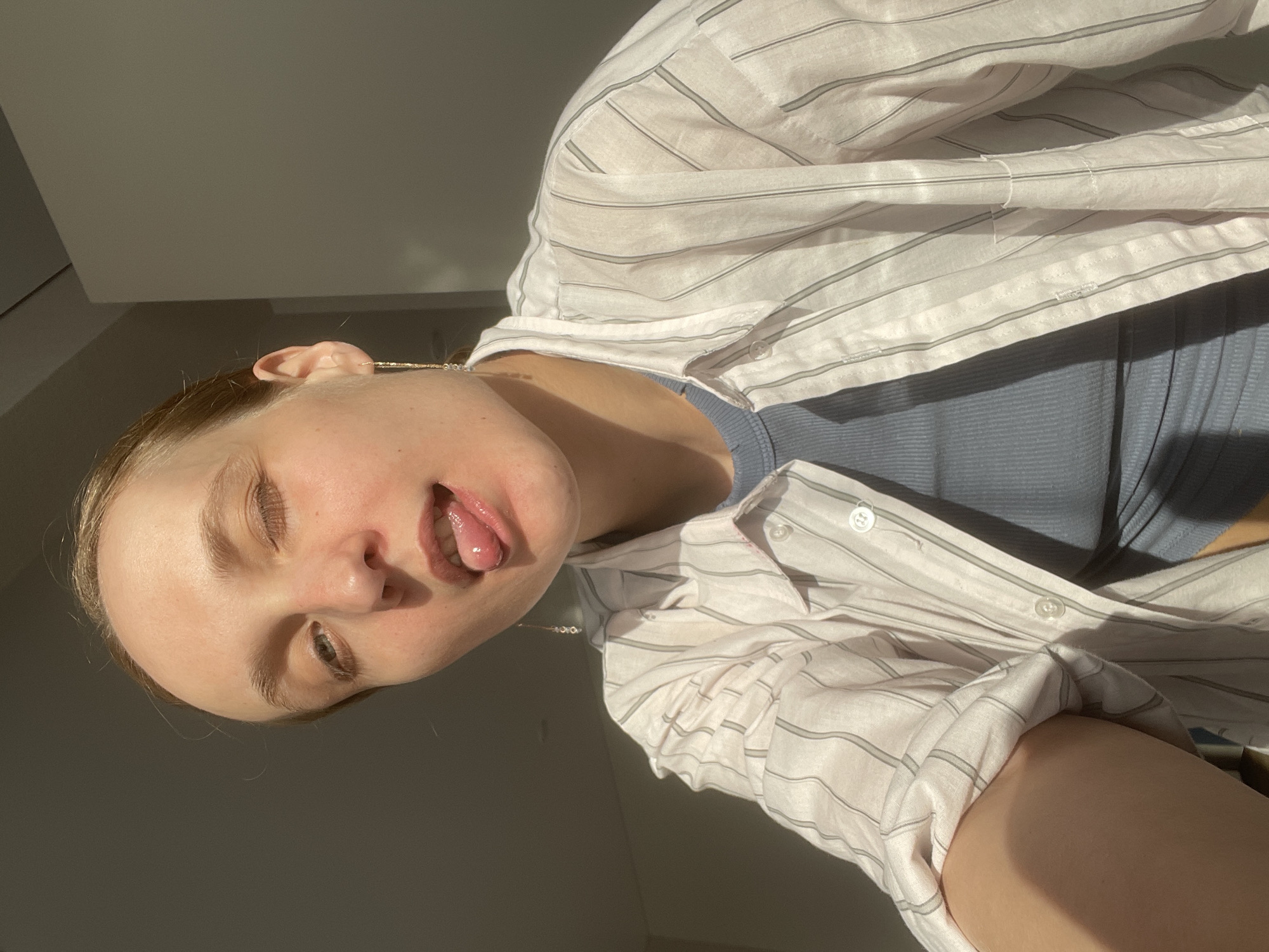 A young, fair-skinned woman is depicted in a close-up, angled shot. Her head is tilted, and she is sticking her tongue out slightly. She has one eye closed and the other partially open. She is wearing a light-colored, striped button-down shirt over a blue top. The background is a mix of light and shadow, suggesting an indoor environment with a window or light source. The photograph may have been taken indoors in Sepych, Russia.