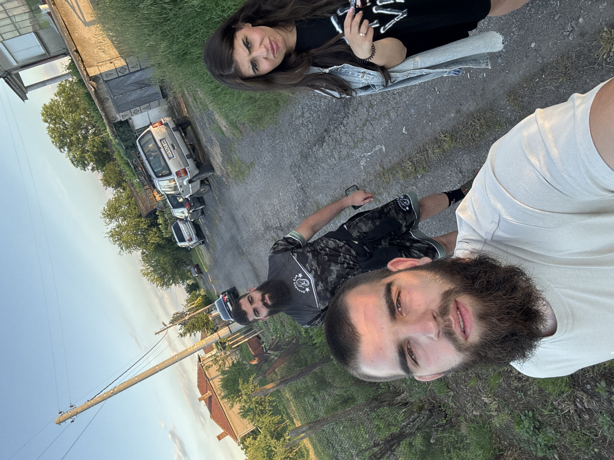 Three individuals stand on a gravel road. A woman with long dark hair holds a phone, wearing a black shirt and denim jacket. Two men with beards are present; one wears a white shirt, the other a dark grey camo shirt. The road is flanked by grass on either side. A series of vehicles, including a white van with the license plate "CH 5826 AH," are parked along the roadside. In the background, there is a building with a red roof, trees, and a utility pole. The location appears to be near a residential area, possibly in Novoselets, Bulgaria.