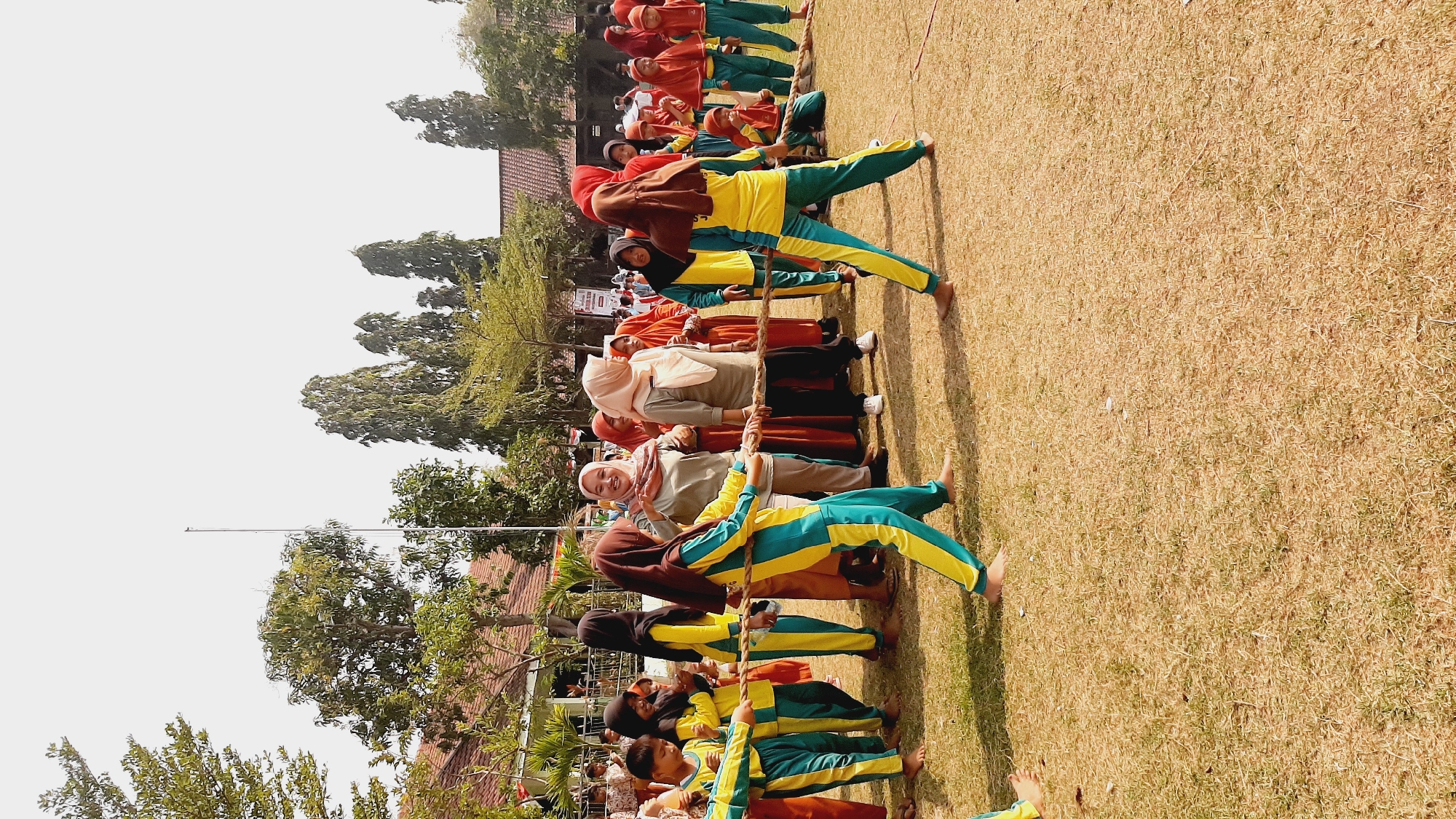 In the city of Sumberjo, Indonesia, a tug-of-war competition is underway on a dry, grassy field. A group of children, predominantly wearing yellow and teal athletic uniforms with headscarves, are engaged in the game, pulling on a thick rope. Several adults, some in headscarves and others in casual attire, are observing and perhaps coaching. In the background, a school building with a red-tiled roof and a flagpole are visible, surrounded by trees. The sky is bright and clear, indicating a sunny day.