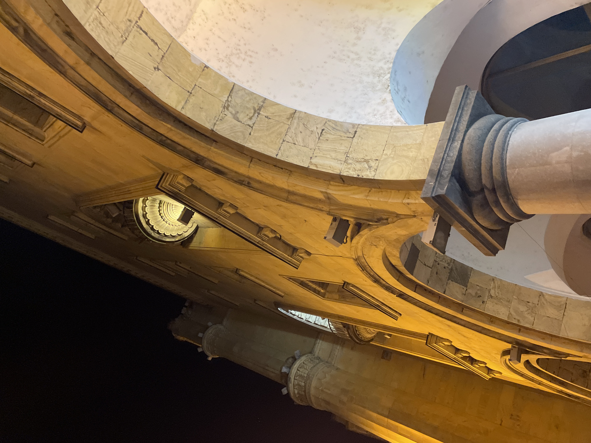 The photograph showcases an interior architectural detail, likely a dome or a high ceiling within a building. The structures are constructed from what appears to be sandstone or a similar light-colored stone. A prominent feature is a series of arches or concentric rings that lead upwards towards a central dome-like structure with a textured surface. Part of a column is visible. The scene is illuminated by artificial lighting, which casts shadows and highlights the textures of the stone. The image is likely located in Tbilisi, Georgia.