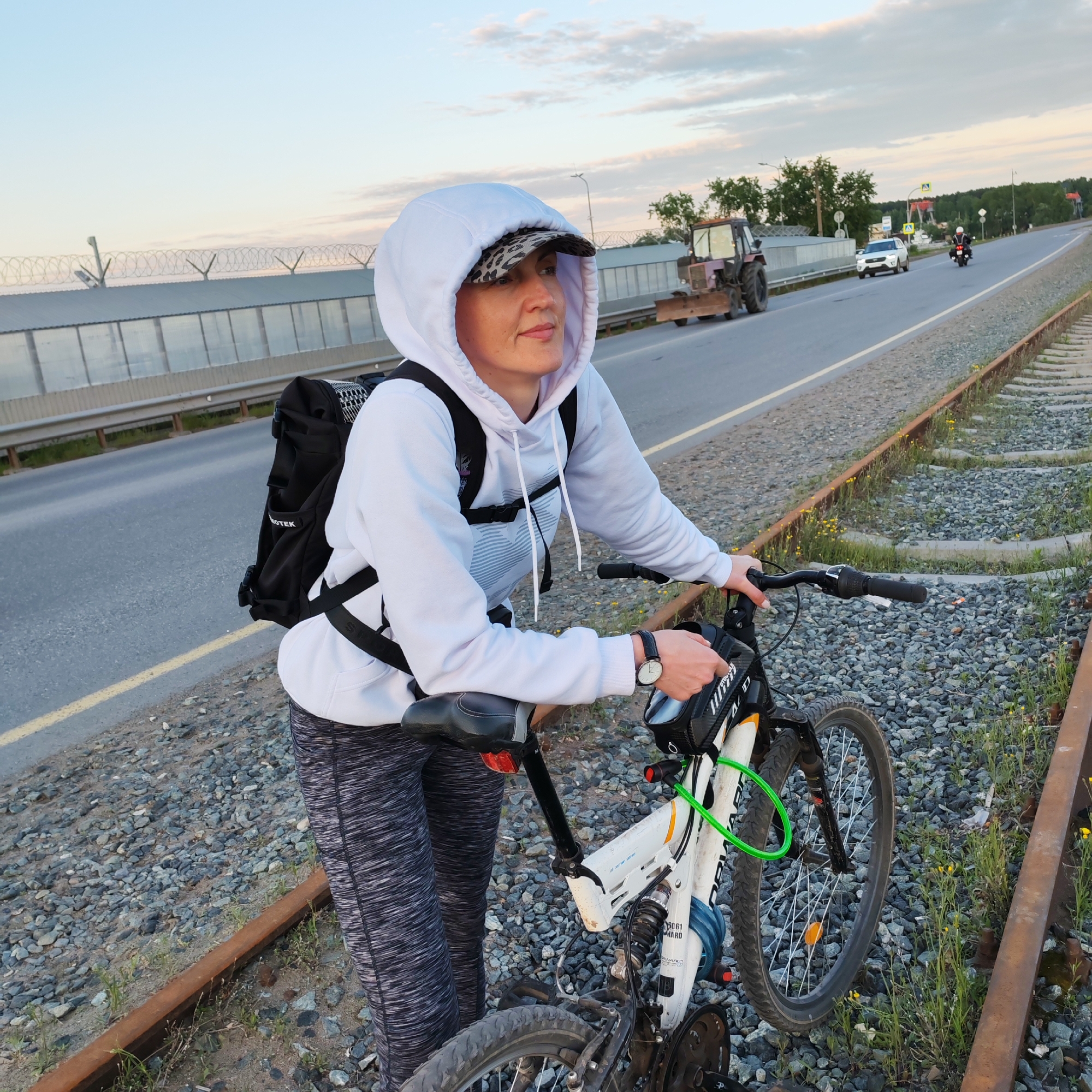 A woman stands next to her white mountain bicycle on railroad tracks, with her hands resting on the handlebars. She is wearing a white hoodie with the hood up over a leopard-print baseball cap, patterned black and grey leggings, and a black backpack with "VOTEK" partially visible. A black watch is on her left wrist, and she looks pensively towards the right. The bicycle has a green cable lock around its frame, and "WARD 5061" is visible on its downtube.
Parallel to the tracks, an asphalt road features a guardrail and a sound barrier topped with barbed wire. Traffic on the road includes a red-brown tractor with a front blade, a white car, and a motorcyclist. Utility poles and trees line the road, with further foliage and hills visible in the distance under a partly cloudy sky. The soft, diffused light suggests late afternoon or early evening. The rusty railroad tracks are surrounded by gravel, green grass, and small yellow wildflowers. The scene is set in or around Chaikovsky, Russia.