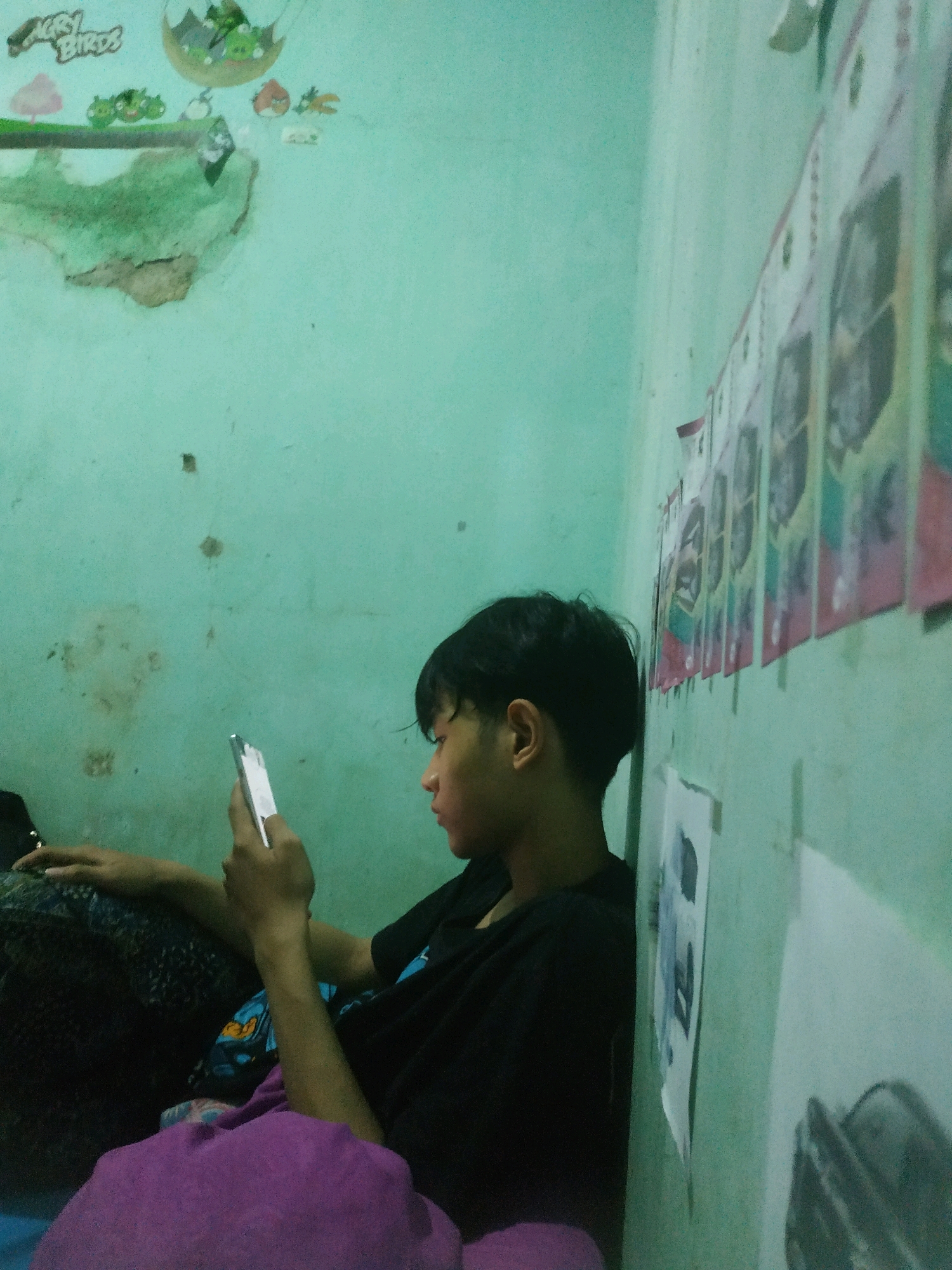 The image depicts a young person sitting on a floor, leaning against a wall, and looking at a smartphone. The person has short, dark hair and is wearing a black t-shirt. They are seated on a dark patterned fabric, with a purple blanket or cloth in the foreground. The room has a light green wall. On the wall to the left, there are some faint cartoon decals, possibly from "Angry Birds," and a section of damaged plaster. On the wall to the right, there are several rows of what appear to be printed images or posters attached to the wall. The scene is dimly lit, creating a somewhat muted color palette. The image appears to be taken indoors, likely in a residential setting in the city of Pacul, Indonesia, given the context.