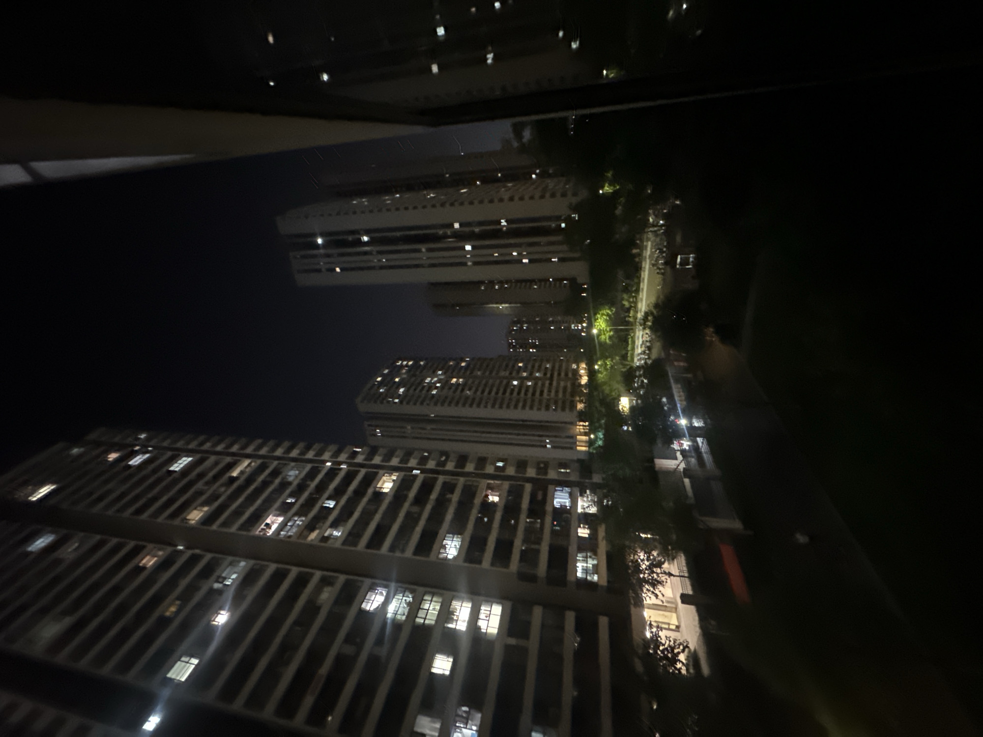 The media file presents a nighttime urban landscape, likely situated in Chongqing High-tech Zone, China, viewed from an elevated perspective. The scene is dominated by several towering, modern high-rise buildings that stretch vertically against a dark, clear night sky.
The architecture of the buildings is characterized by repetitive patterns of windows and balconies, with many individual units displaying illuminated interiors. These warm lights punctuate the dark facades, indicating ongoing activity or occupancy within the residential or commercial structures. The foreground and sides of the image show indistinct dark structures, possibly a balcony or window frame from which the photograph was taken.
At ground level, between the prominent high-rises, a network of illuminated streets or pathways is visible. Trees with dense foliage line these routes, their forms subtly highlighted by artificial streetlights. Faint glints of light suggest distant vehicles or other urban elements, contributing to the sense of a vast, active city.
The overall atmosphere is one of quiet urban life at night. There are no discernible people or specific events captured. The image showcases the dense development and verticality typical of major Chinese cities, emphasizing artificial light as the primary source of illumination against the backdrop of night. No discernible text is visible within the frame.