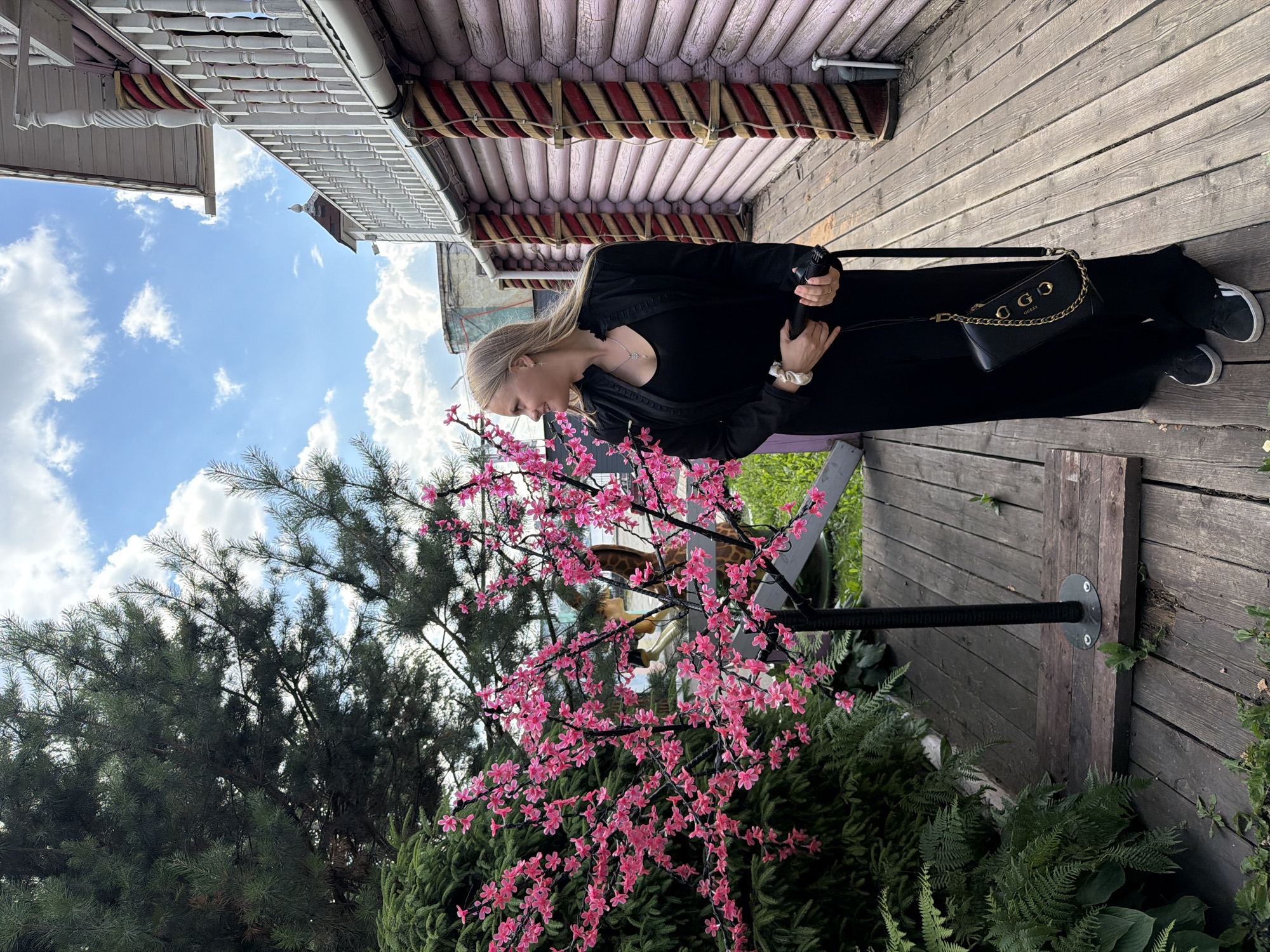 A young blonde woman, dressed in a long black dress and black sneakers, stands on a wooden deck. She is holding a black object in her right hand, possibly a phone, and a black handbag with a gold chain is slung over her left shoulder. She is looking down with a neutral expression. In front of her is a small artificial tree with bright pink blossoms. Behind her, part of a wooden building with pink and red decorative elements is visible, along with a glimpse of a blue sky with white clouds. The scene appears to be outdoors, possibly in a garden or park, with lush green foliage in the foreground and background. The lighting suggests it is daytime. The wooden structures and architectural details hint at a location in Russia, consistent with the provided context of Izhevsk. There is no visible text in the image.