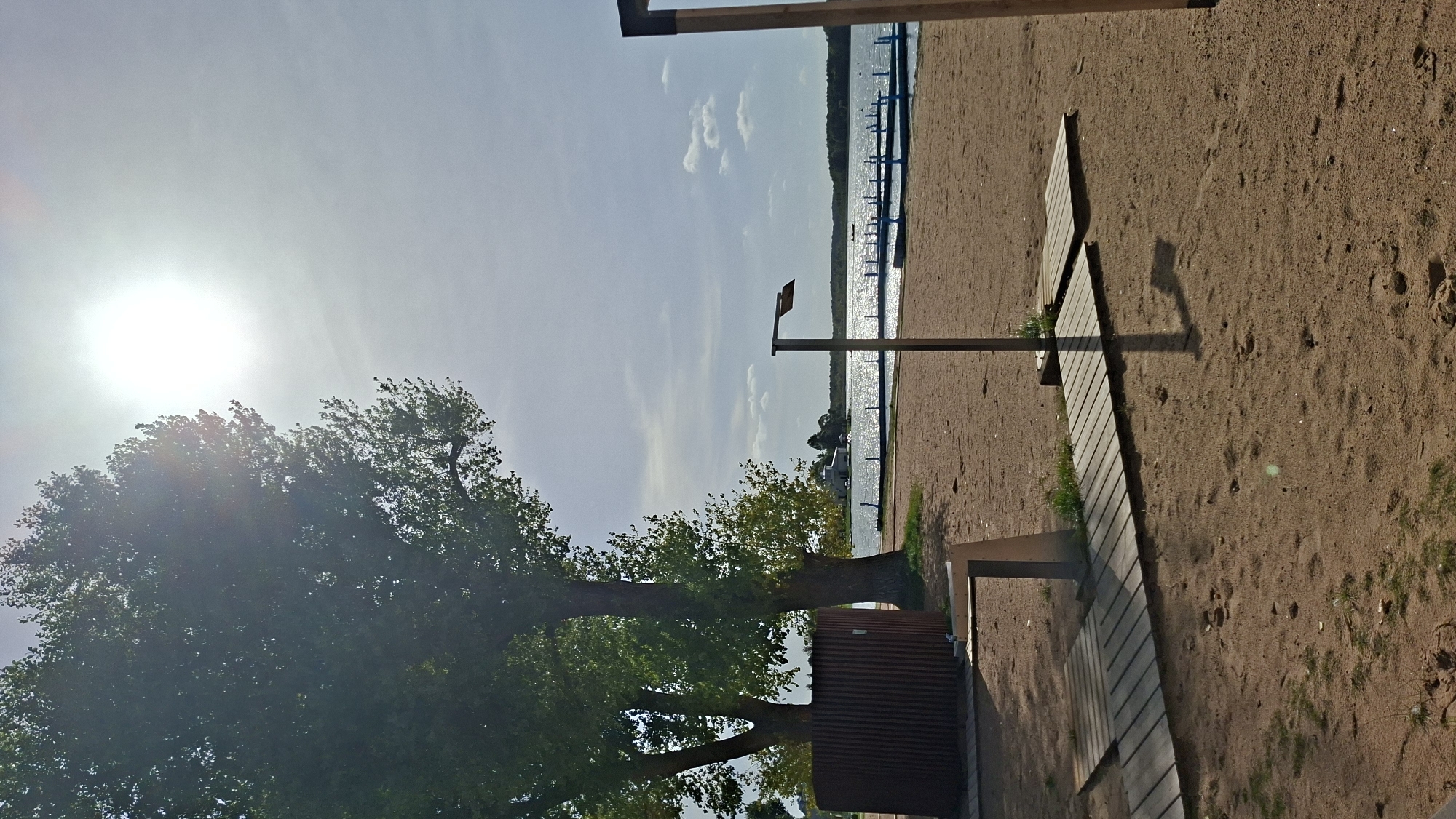 The image depicts a sandy beach area next to a body of water, likely Lake Myadel, in Myadzyel, Belarus. The sun is bright in the sky, casting a glare and shadows. On the sand, there are wooden walkways leading towards the water. A shower structure is visible on the left, with a corrugated metal enclosure. In the distance, across the water, there are buildings and what appears to be a dock or pier. Several trees with green foliage are situated on the left side of the image. The water is calm and reflects the sunlight, and a few clouds are present in the sky. The scene conveys a sense of a recreational area, likely a public beach, on a sunny day.