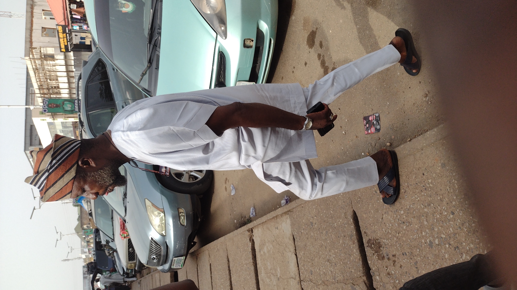 An image taken in the city of Jos, Nigeria, shows a man in traditional attire standing next to a curb, facing left. The man is wearing a striped cap, a white striped shirt, white pants, and sandals. He is holding a black cell phone. The background features several cars parked along the street, and buildings can be seen in the distance. The scene is outdoors with sunlight and street lighting.