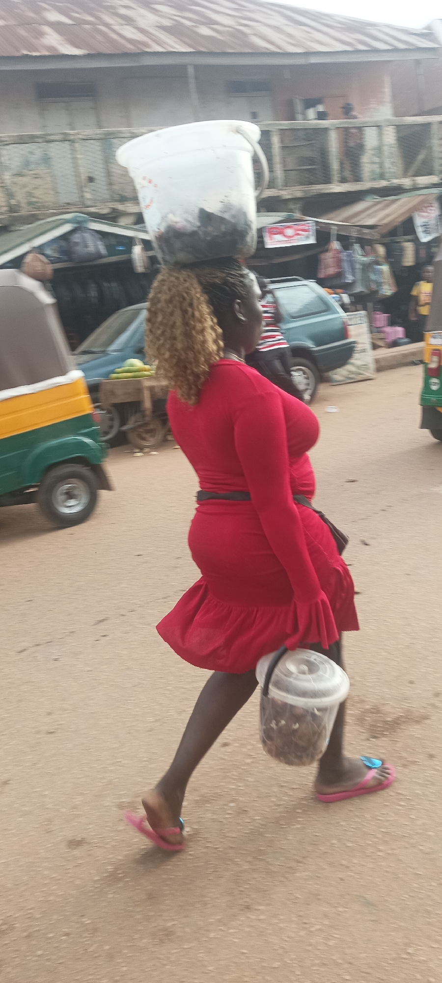 A forensic examination of the image depicts an outdoor scene in Bukuru, Nigeria, featuring a woman walking on a light brown, unpaved road. The woman, positioned centrally and facing left, wears a long-sleeved red dress with a ruffled hem, a black belt, and dark leggings or stockings. Her hair is styled in a blonde curly ponytail over dark braided roots. She wears pink flip-flops on both feet.
She is engaged in carrying two containers: a large white plastic bucket filled with dark, granular or lumpy material is balanced on her head, and a smaller transparent plastic bucket with a white lid, containing similar dark material, is held in her right hand at her side.
The background reveals a street environment consistent with a market or commercial area. To the left, a green and yellow three-wheeled auto-rickshaw is partially visible. Further back, a dark green hatchback car is parked or moving slowly. Behind the car, a structure with a corrugated metal roof and an upper-level balcony or open area is visible. Market stalls with various items, including bags and what appear to be clothing, hang for display. A white sign with "POS" in red lettering is affixed to a wooden frame in the mid-ground. Other individuals are partially visible: one person in a striped top behind the dark green car, and another in a yellow shirt on the far right. The overall impression is a busy street scene.