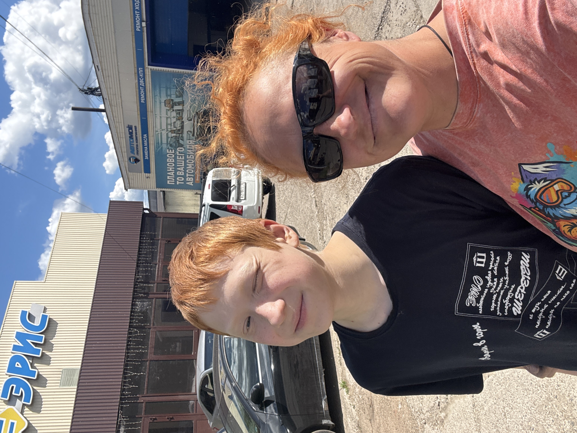 A selfie, slightly rotated, shows a woman with red, curly hair wearing sunglasses and a coral-colored shirt with a graphic design on the right shoulder, standing next to a younger red-haired boy in a black t-shirt with text in Russian. The background indicates they are near a building with a sign that reads "ЭРИС" in large, blue letters. Further behind them is another building with a sign mentioning car maintenance and oil changes: "АВТОМОЁ ГРУППА КОМПАНИЕ ЗАМЕНА МАСЛА ПЛАНОВОЕ ТО ВАШЕГО АВТОМОБИЛЯ РЕМОНТ ДВС-КПП РЕМОНТ ХОДОВОЙ". A white van with the license plate "A278CC" is also visible. Based on the "ЭРИС" sign, the location can be identified as the city of Chaikovsky, Russia.